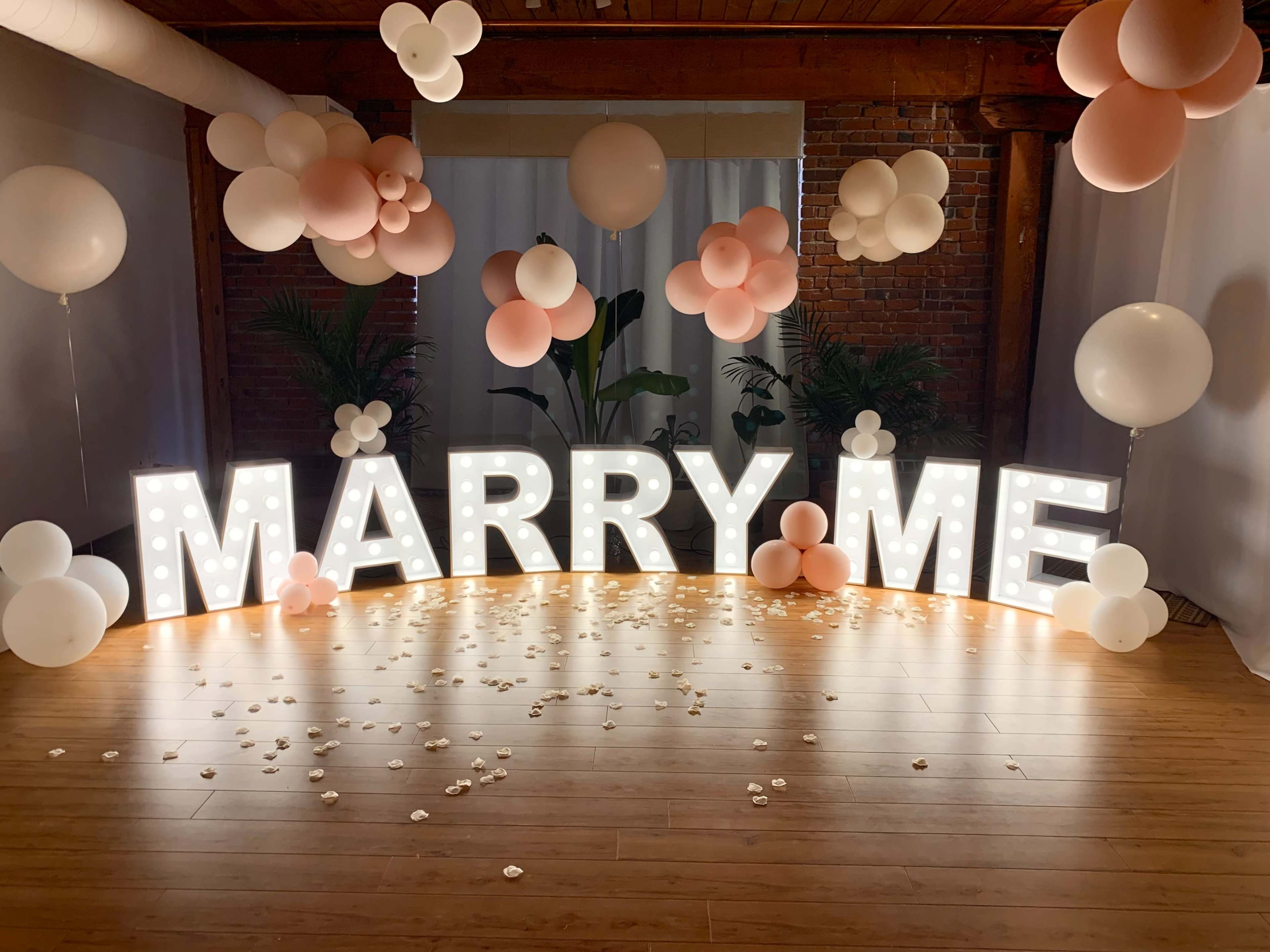 A decorative setup with a large illuminated "MARRY ME" sign surrounded by balloons and floral accents on a wooden floor.