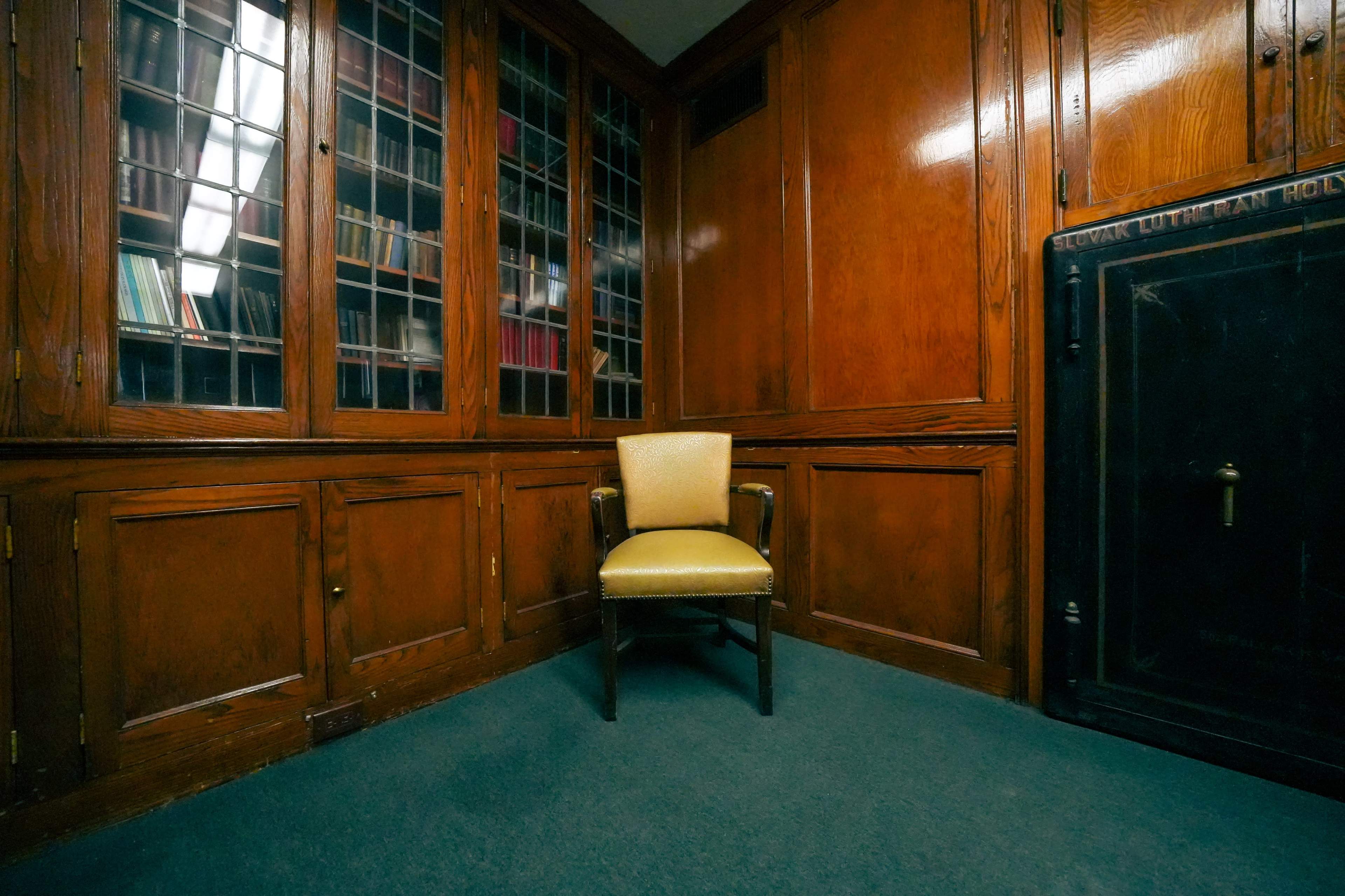 The image shows a wooden-paneled room with a single yellow chair positioned beside a dark safe and a windowed bookshelf filled with books.