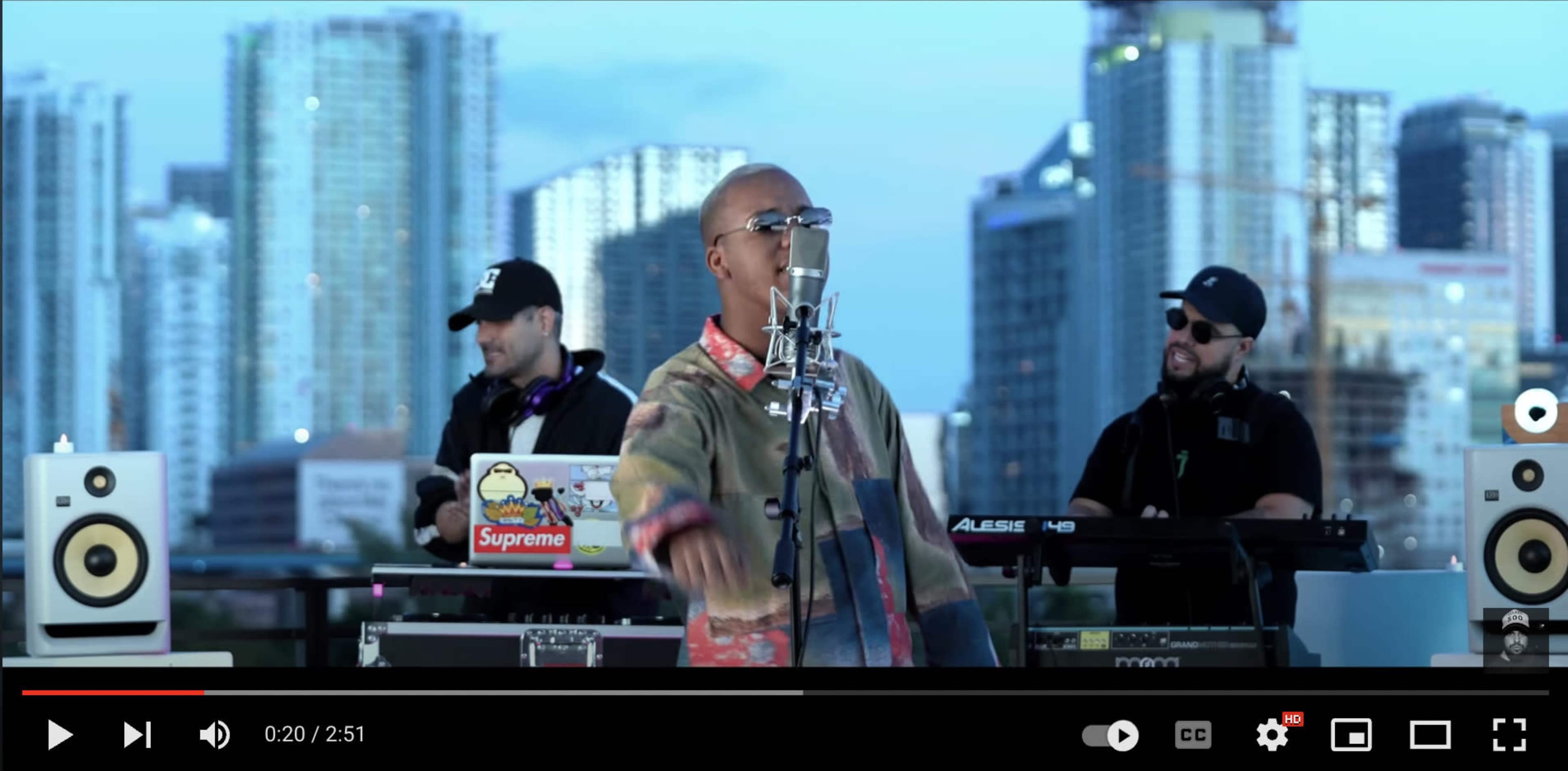 A vocalist sings into a microphone while two musicians play instruments in an urban setting with skyscrapers in the background.