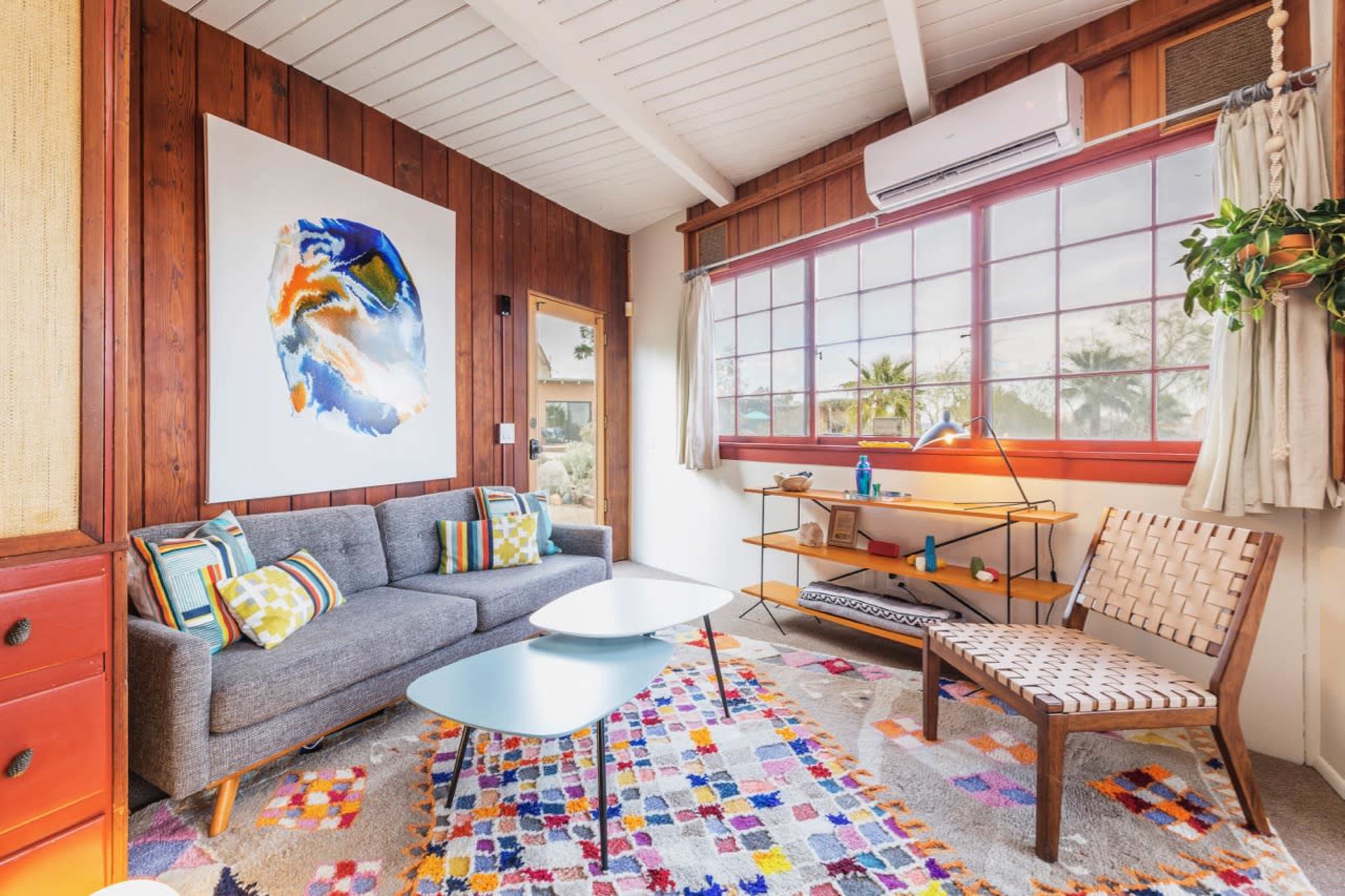 A cozy living room featuring a gray sofa with colorful cushions, a light blue coffee table, and a patterned rug, along with wooden walls and a large window.