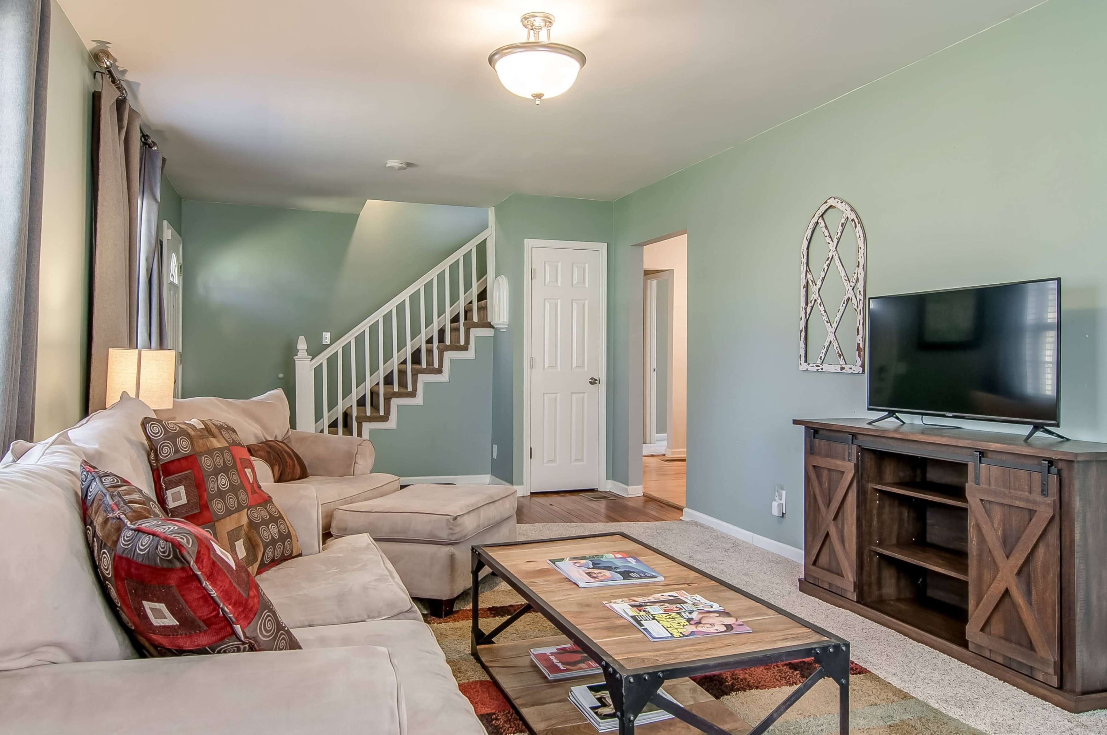 A cozy living room features a beige couch, a wooden coffee table with magazines, and a TV stand against a light green wall, with a staircase leading to an upper level.