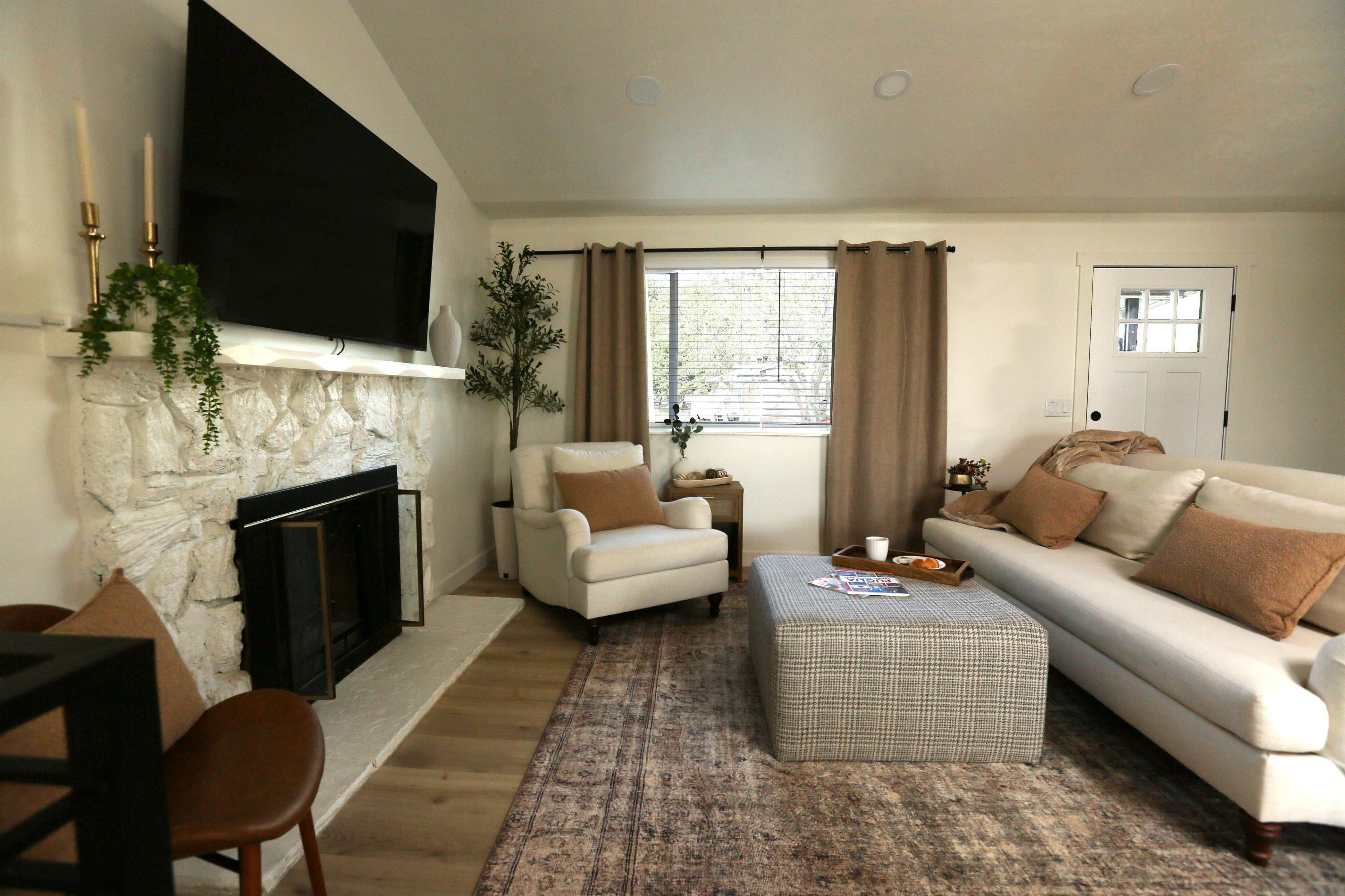 The image shows a modern living room featuring a white sofa, a patterned ottoman, a stone fireplace, and a large flat-screen TV mounted above it.