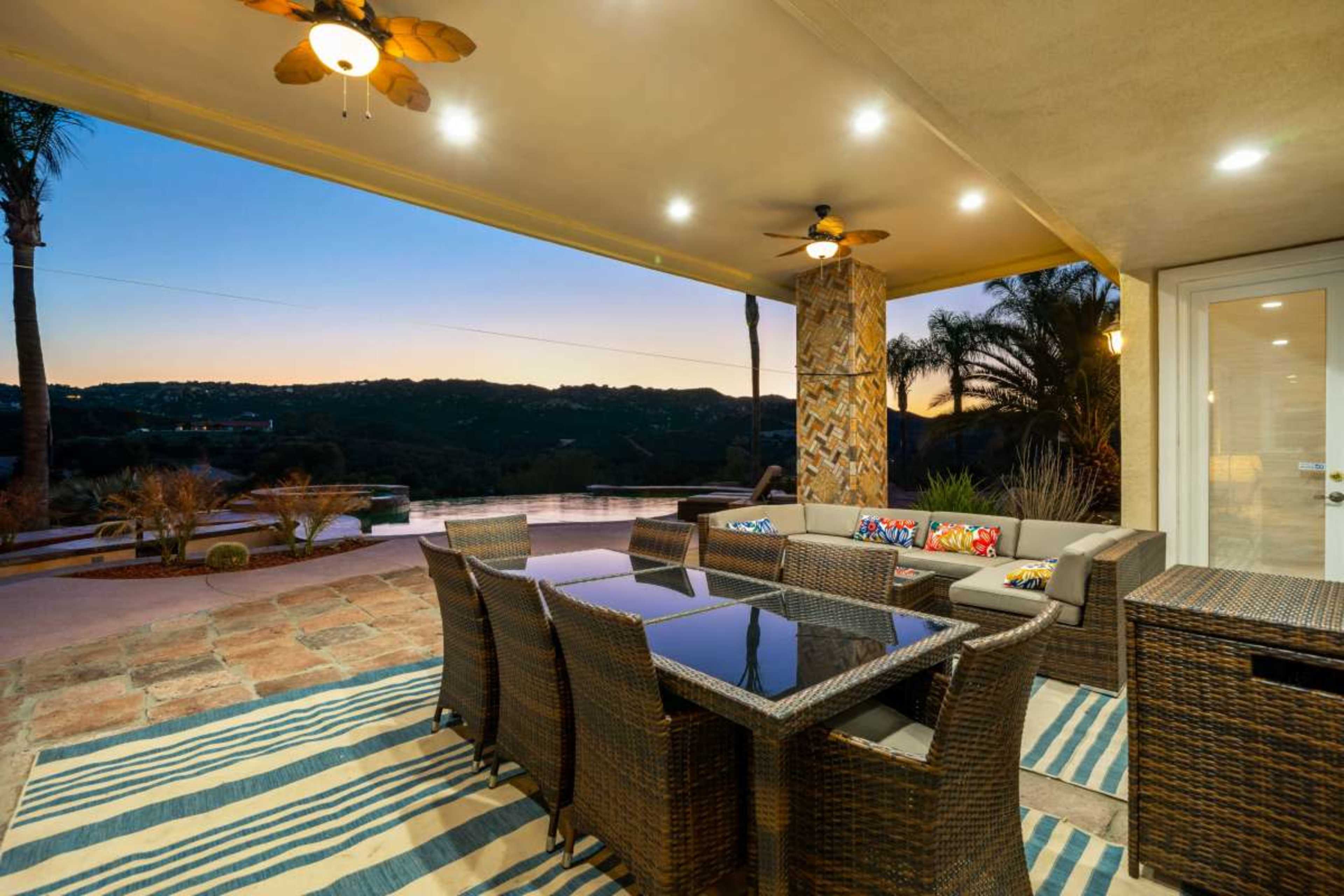 A patio area features a large dining table and chairs with a view of the surrounding landscape during sunset.
