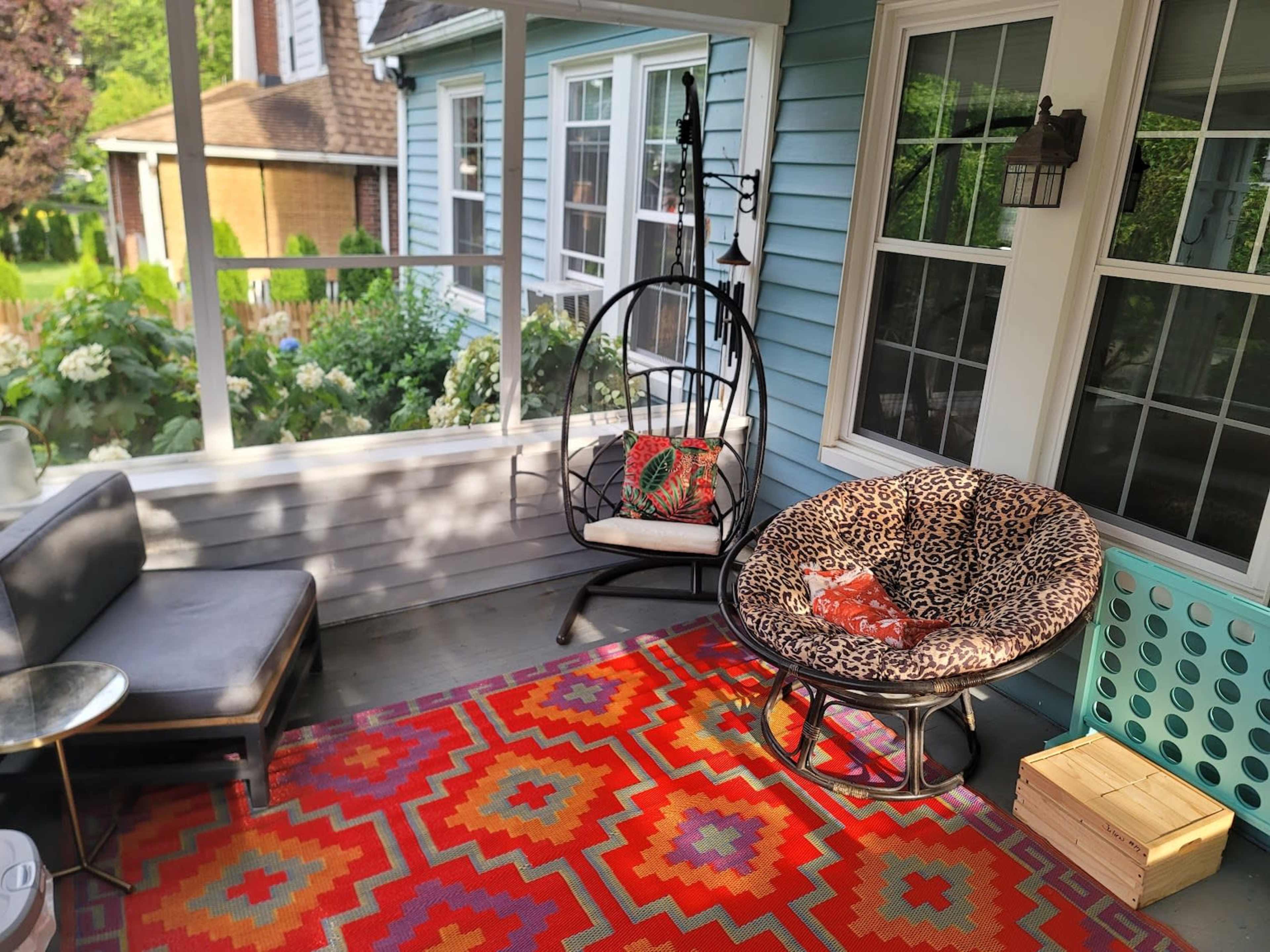 A screened porch with a colorful geometric rug, a hanging chair, a round chair with a leopard print cushion, and a small side table.