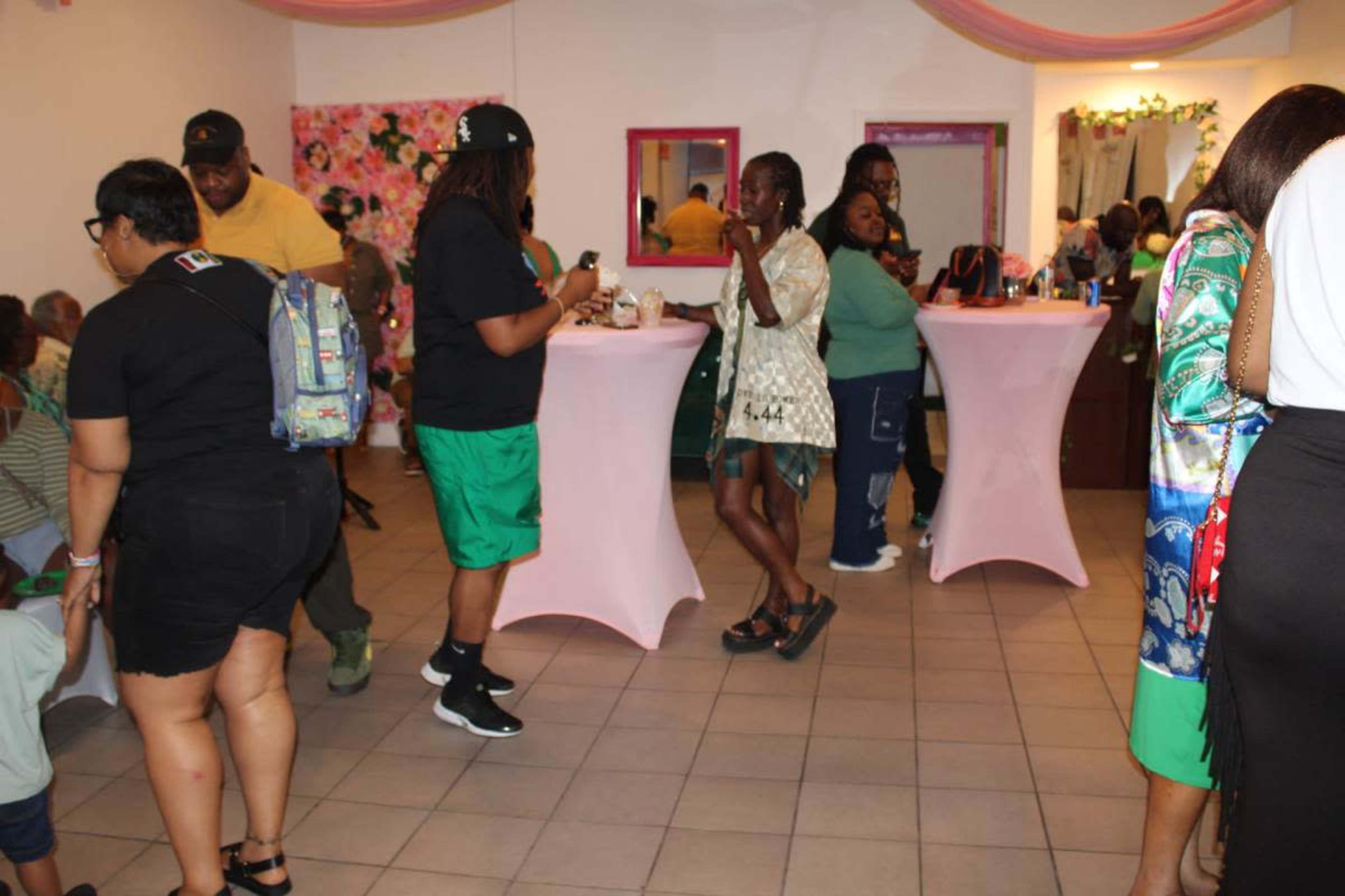 A group of people socialize at a festive indoor event featuring pink tablecloths and floral decorations.
