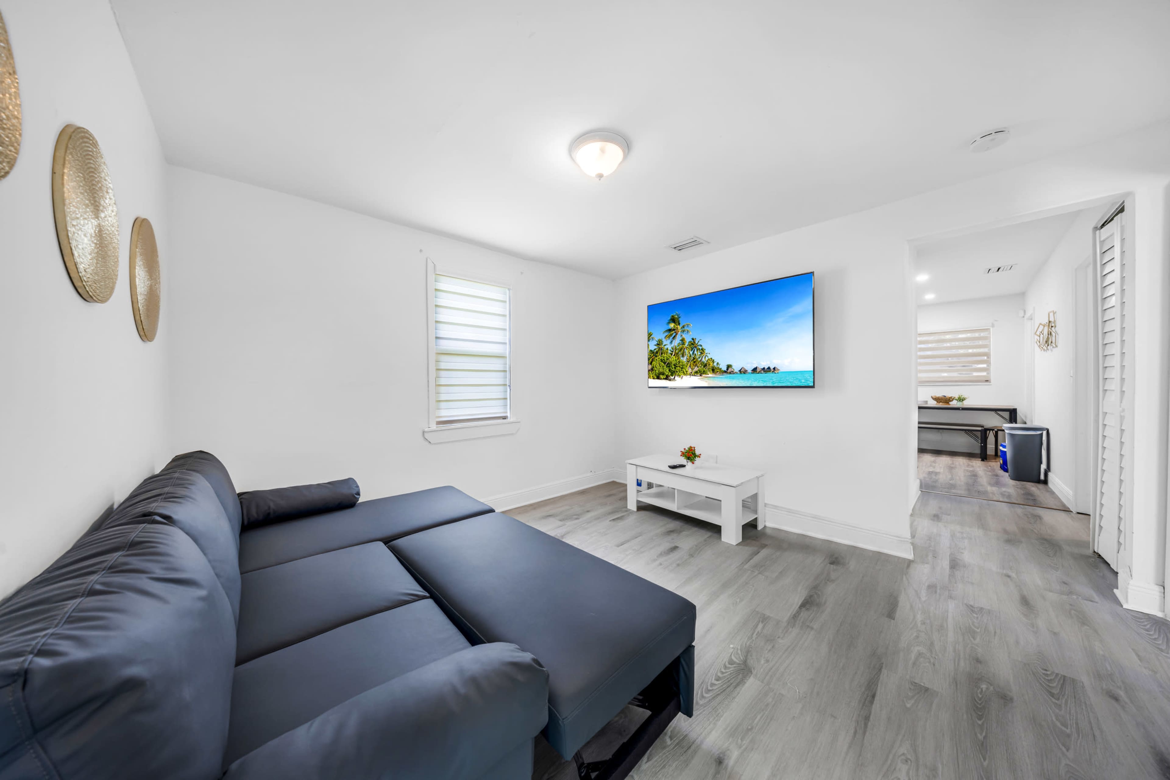 The image shows a modern living room featuring a dark sectional couch, a white coffee table, and a large wall-mounted TV displaying a beach scene.