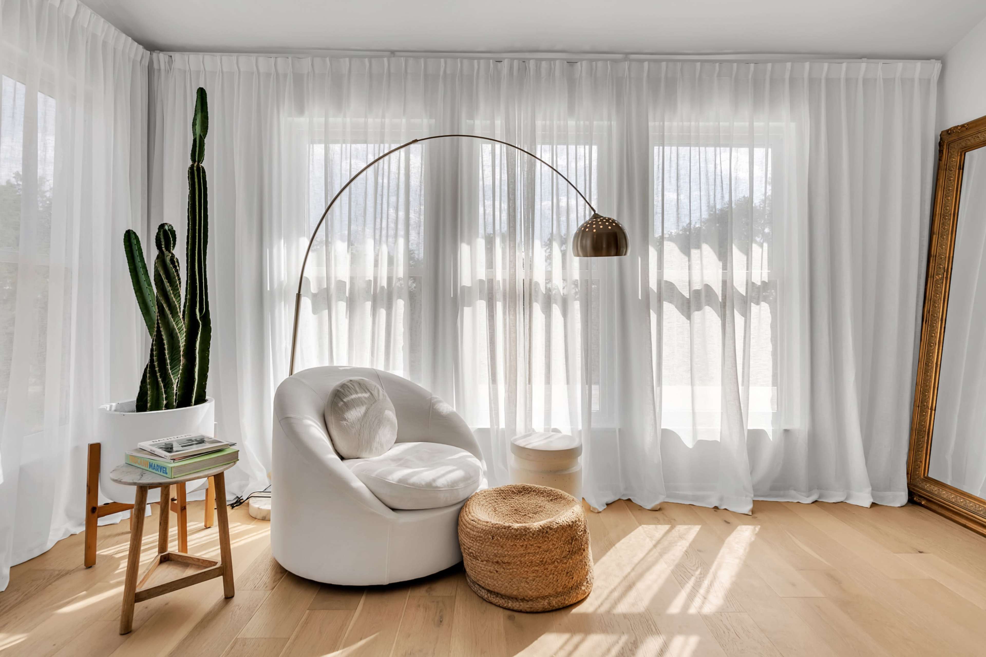 A white armchair and a round ottoman are positioned near large windows with sheer curtains, alongside a tall cactus and a floor lamp.