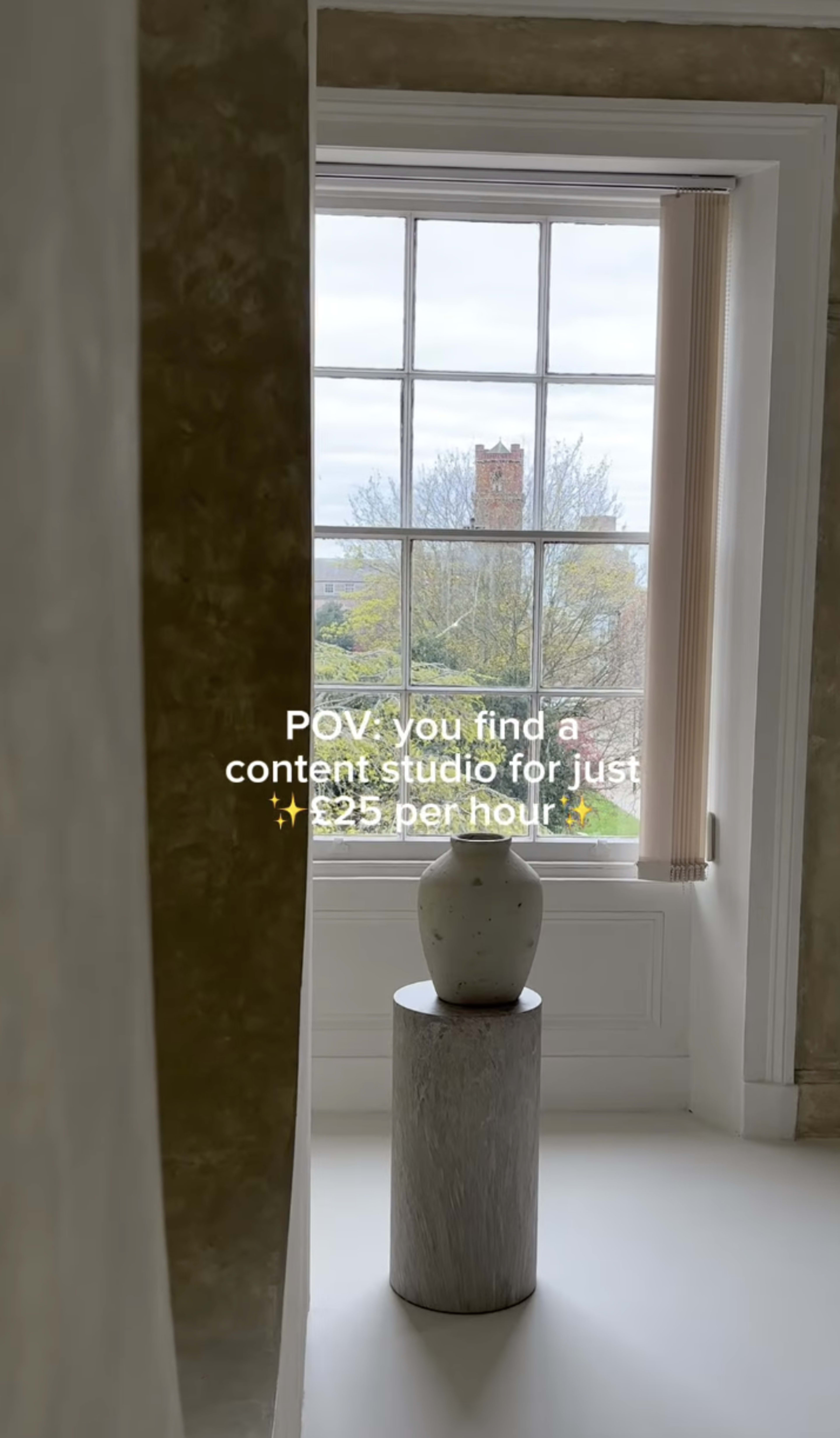 The image shows a content studio with a window view, featuring a cylindrical pedestal with a vase on top.