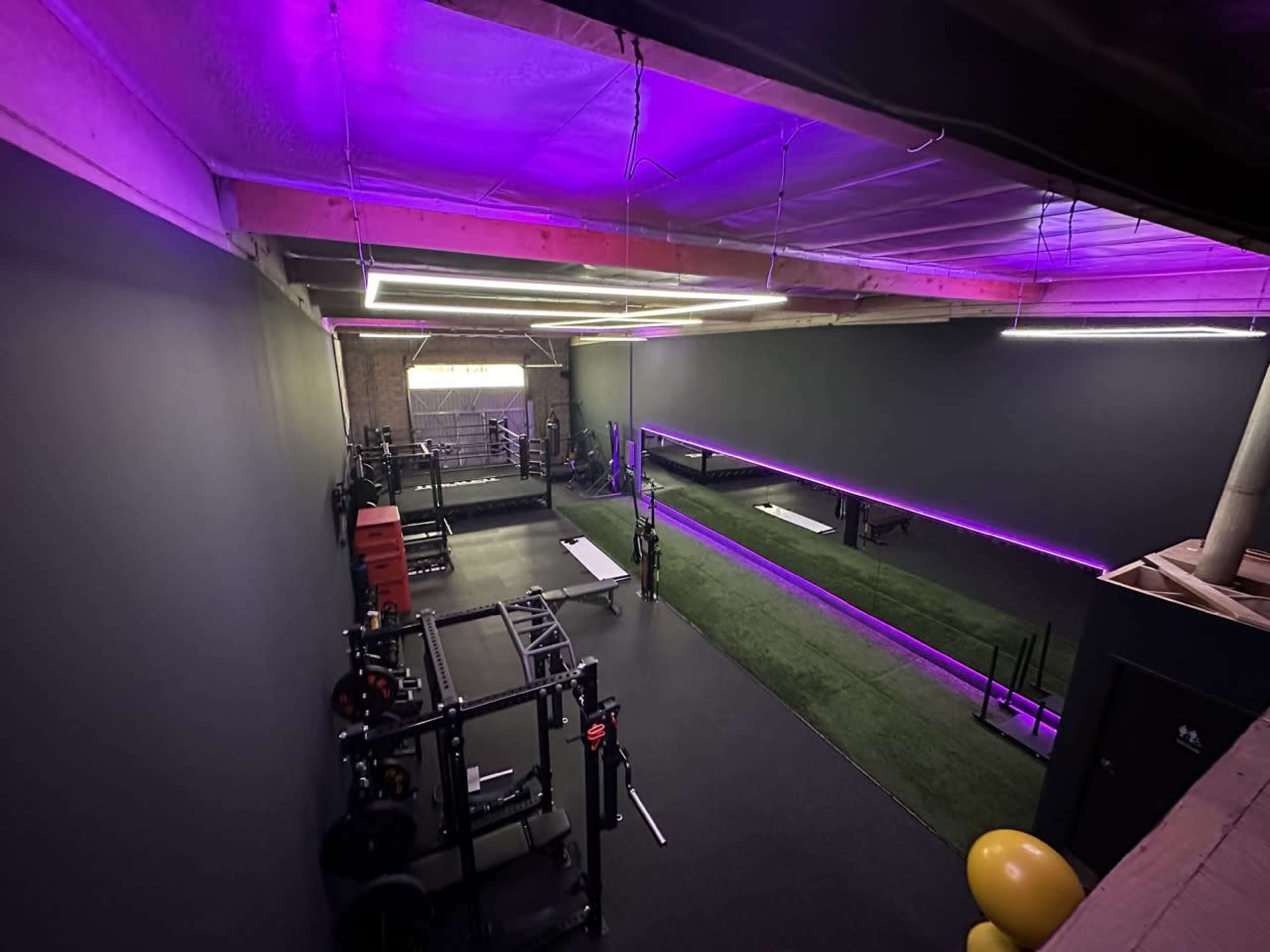 The image shows a modern gym with dark walls, purple lighting, and various workout equipment arranged along the floor.