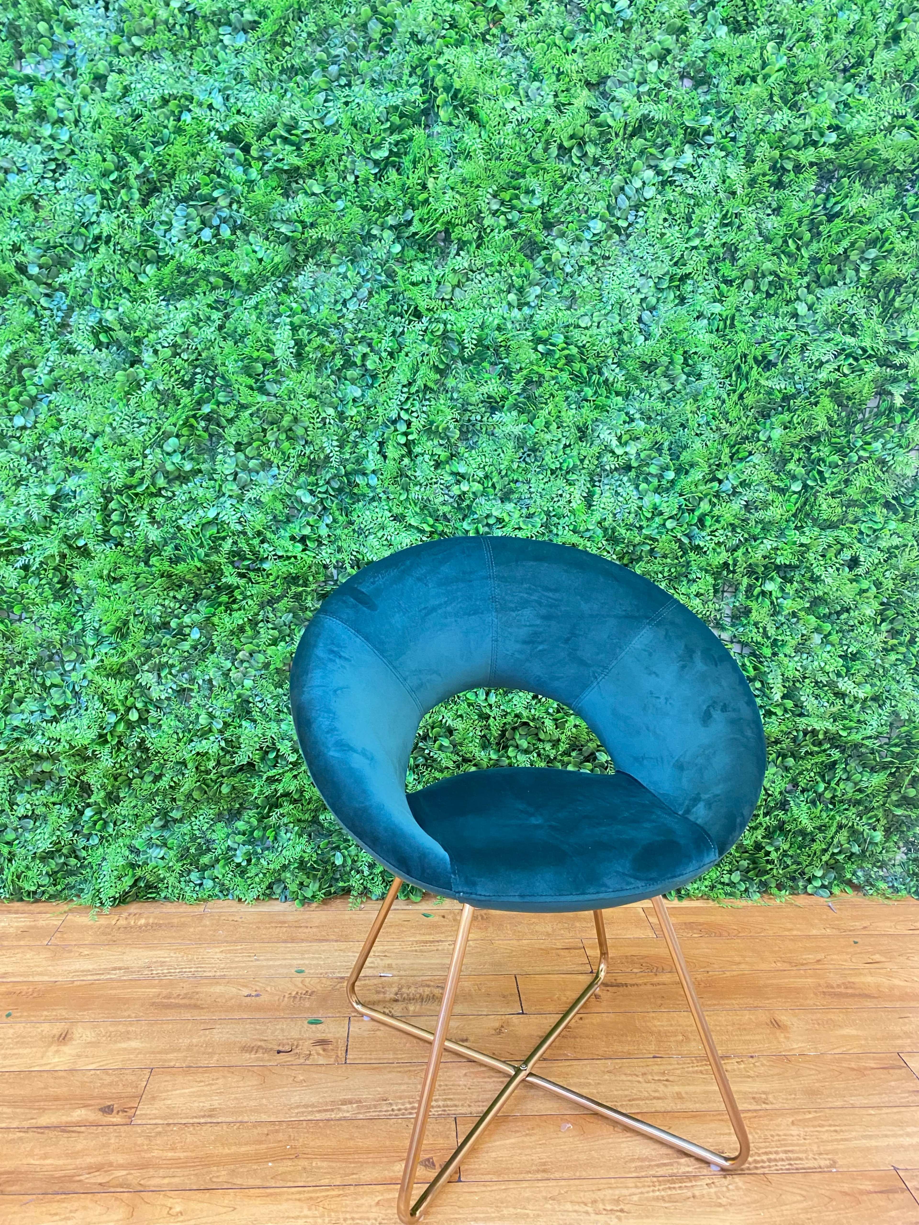 A teal velvet chair with a circular design is positioned against a lush green wall of artificial foliage.
