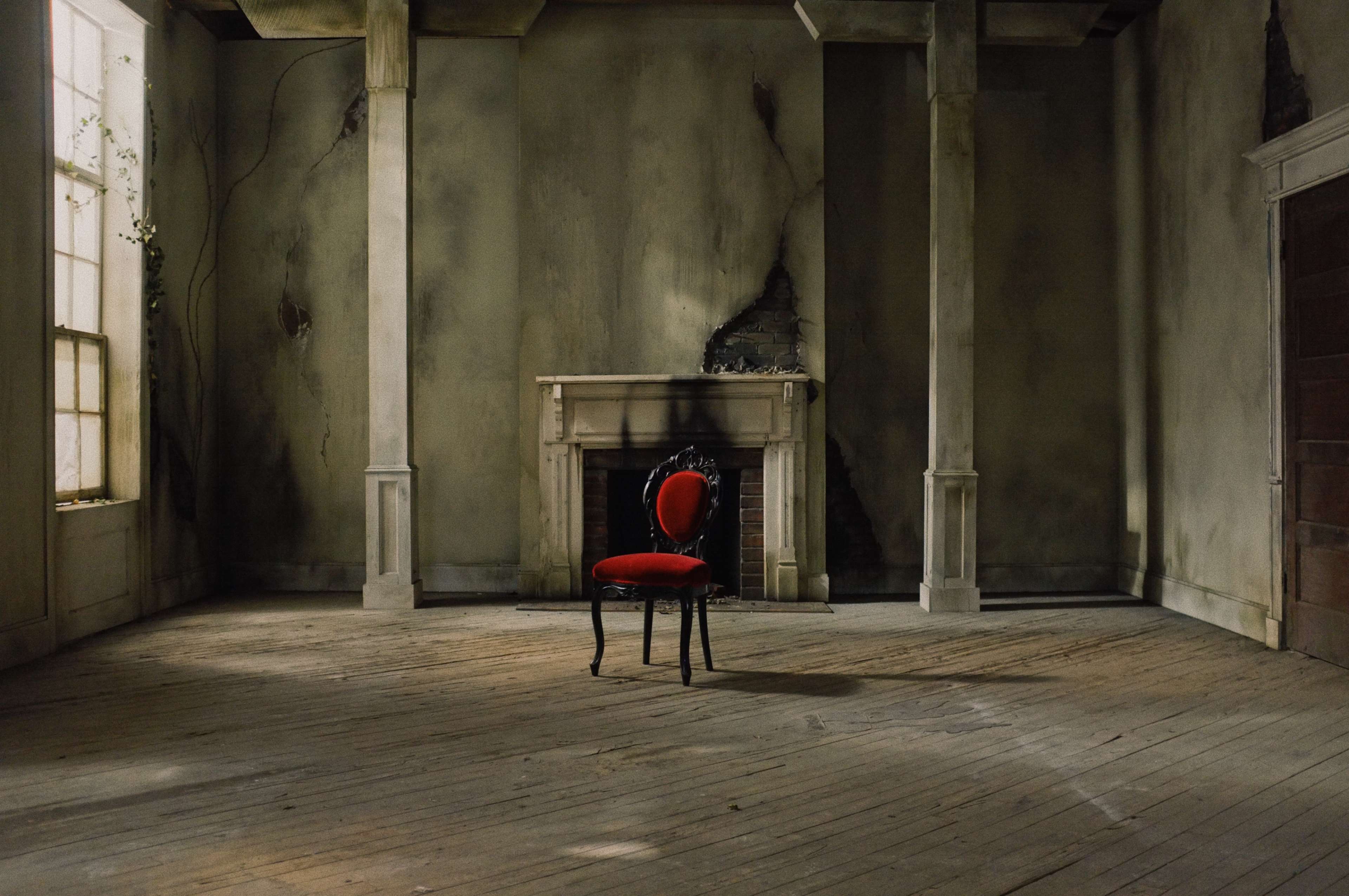 An empty room features a single red chair in front of a fireplace, with peeling walls and wooden floors.