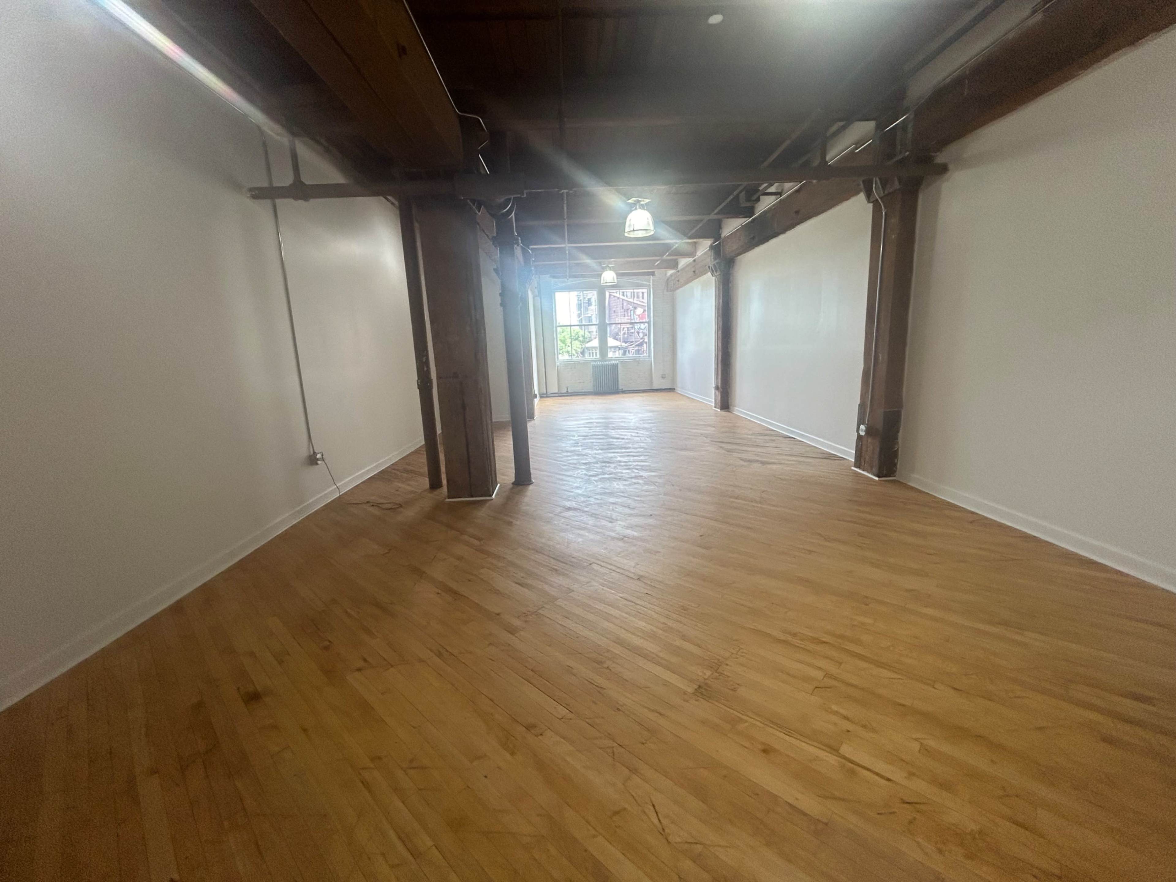 A spacious room with hardwood flooring and exposed wooden beams, illuminated by natural light from windows at the far end.