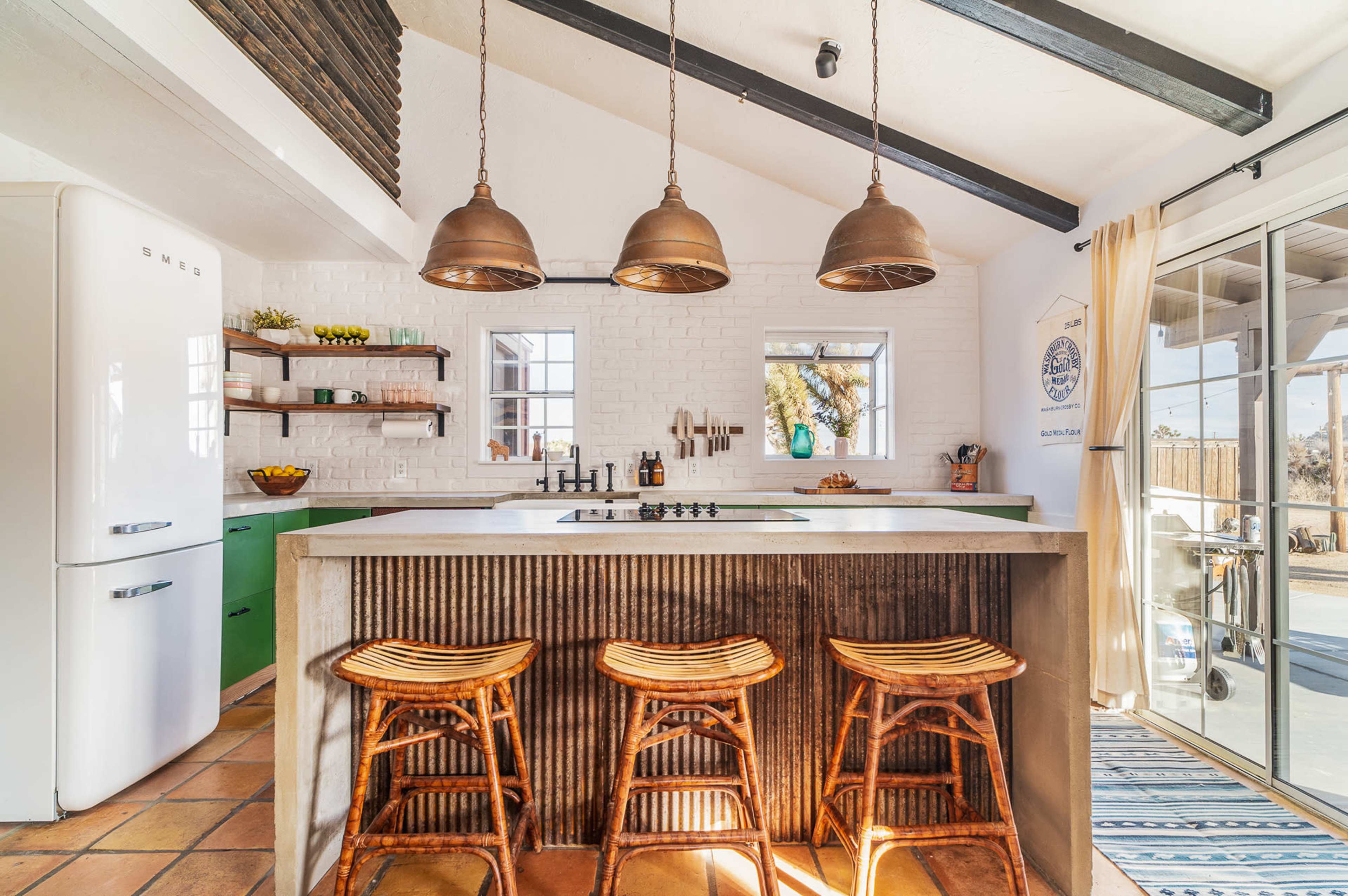 A modern kitchen features a concrete island with three wooden stools, pendant lights, and large windows overlooking an outdoor area.