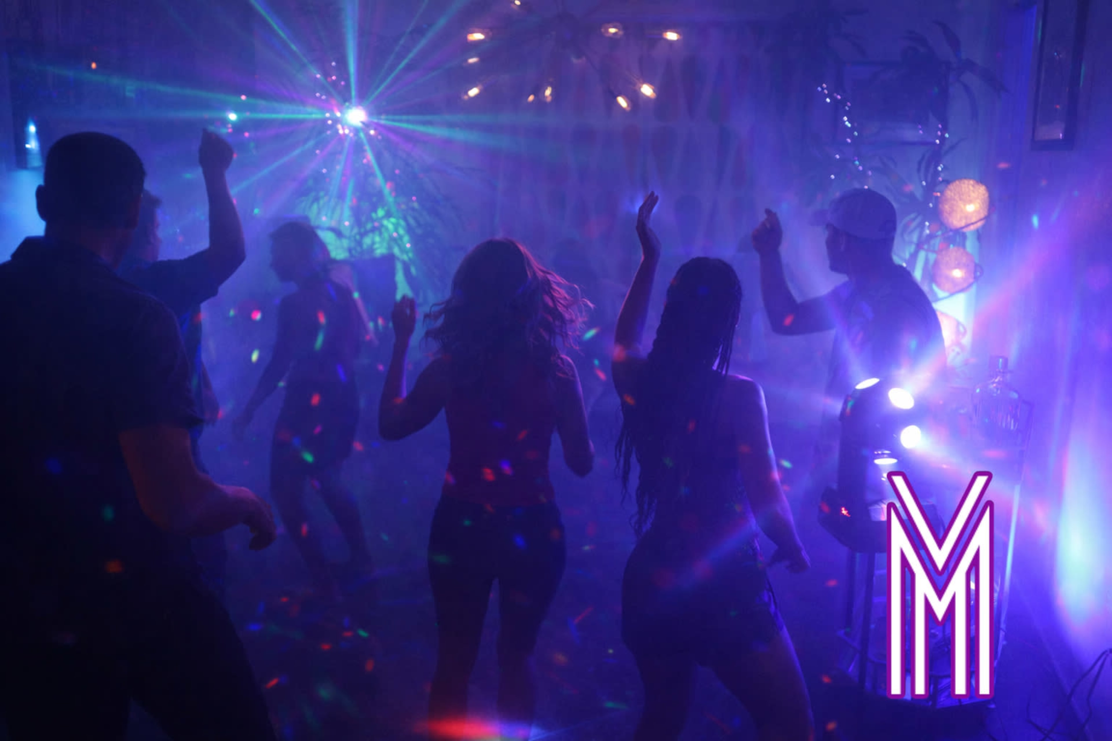 A group of people dance under colorful lights in a dimly lit room filled with smoke and decorations.