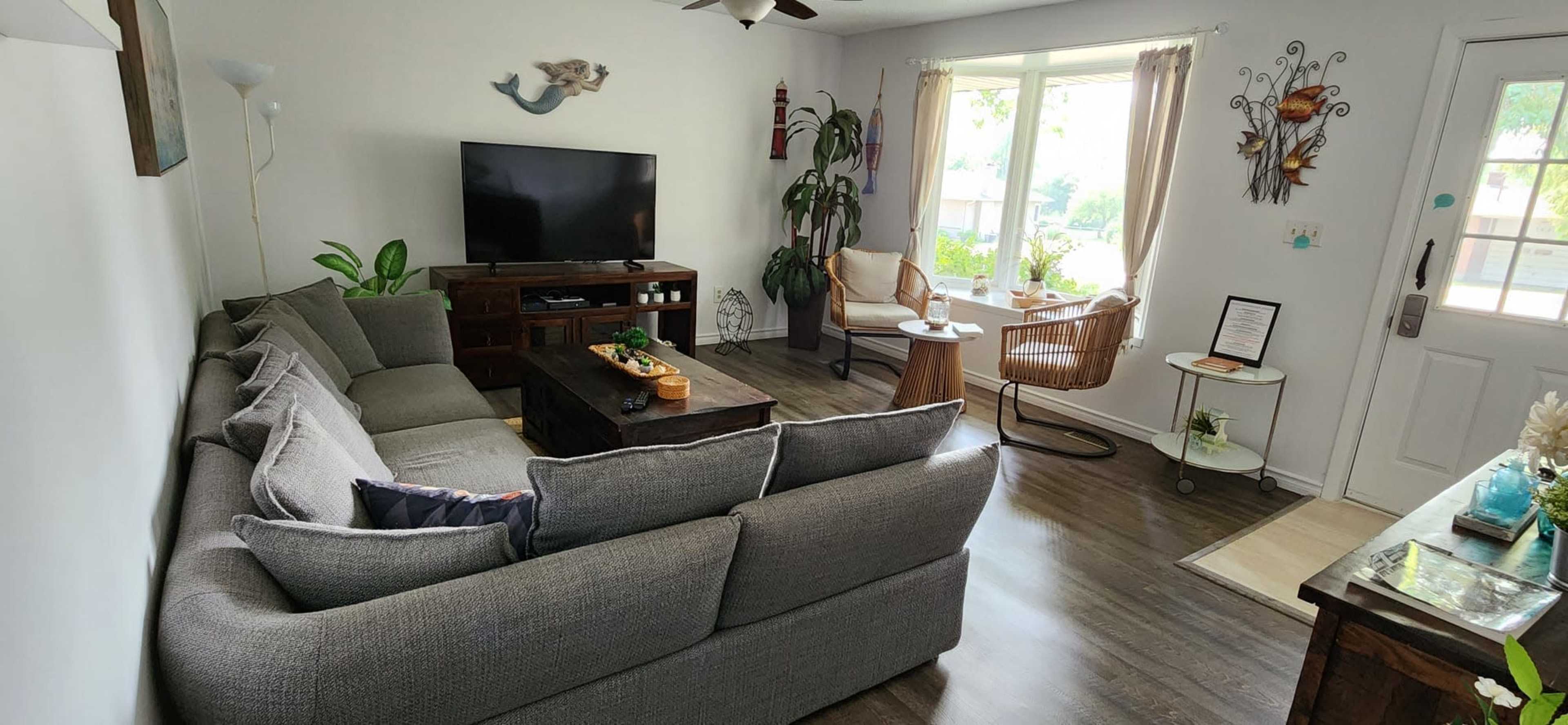A living room features a gray sectional sofa, a TV stand with a television, a coffee table with decorative items, and seating by a window with plants and wall decorations.