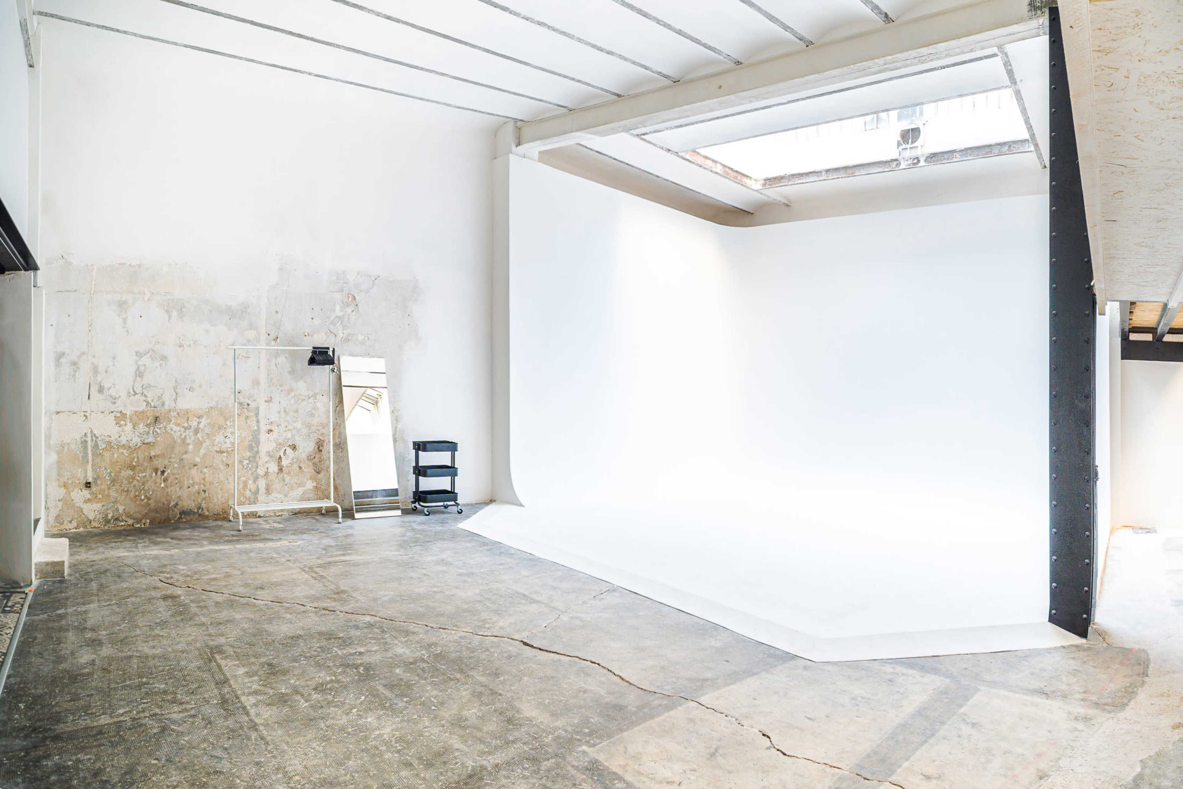 The image shows a spacious, bare studio with a white backdrop, a concrete floor, and minimal furnishings, including a mirror and a black shelving unit.