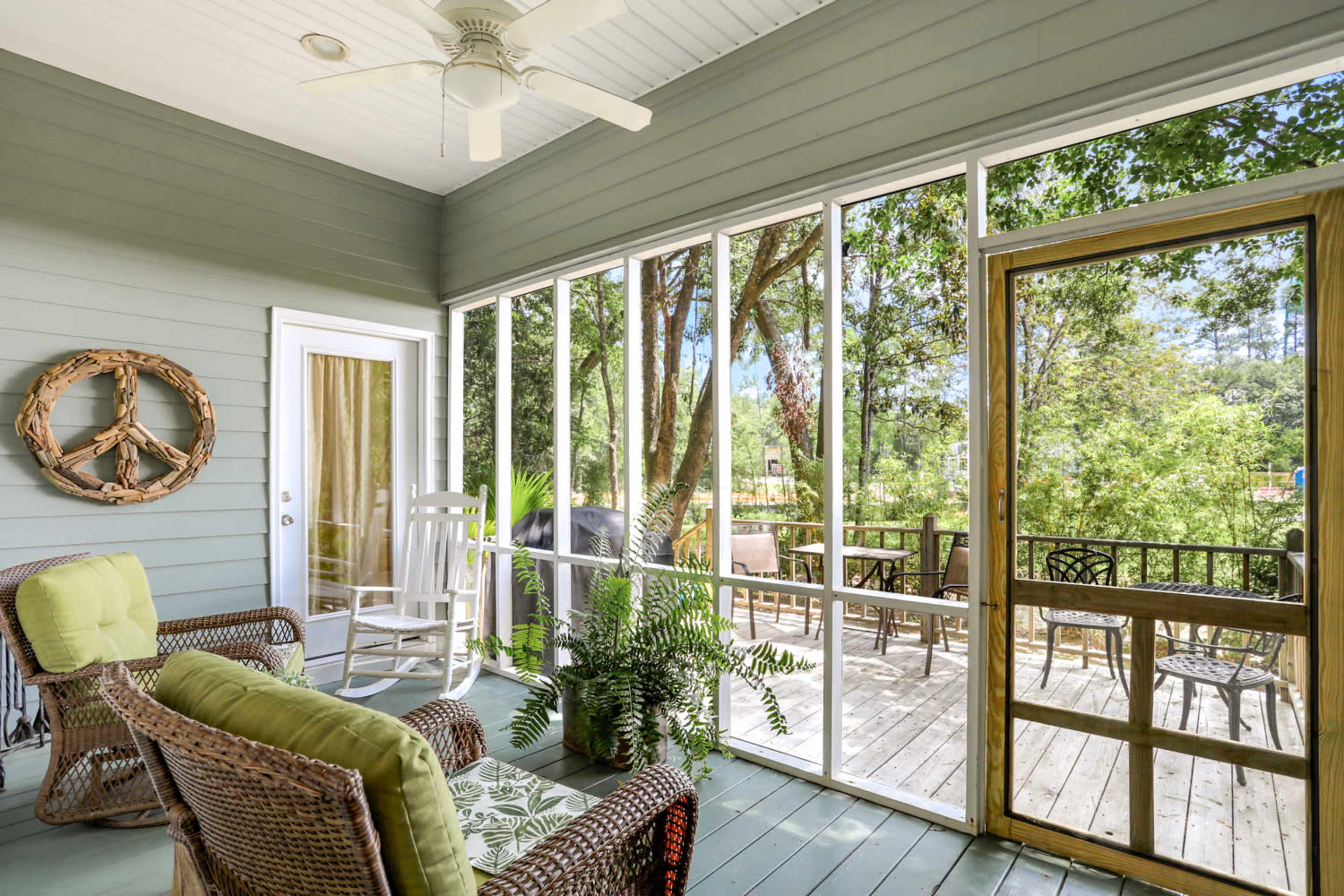 A screened porch features wicker furniture and greenery, overlooking a deck and trees.