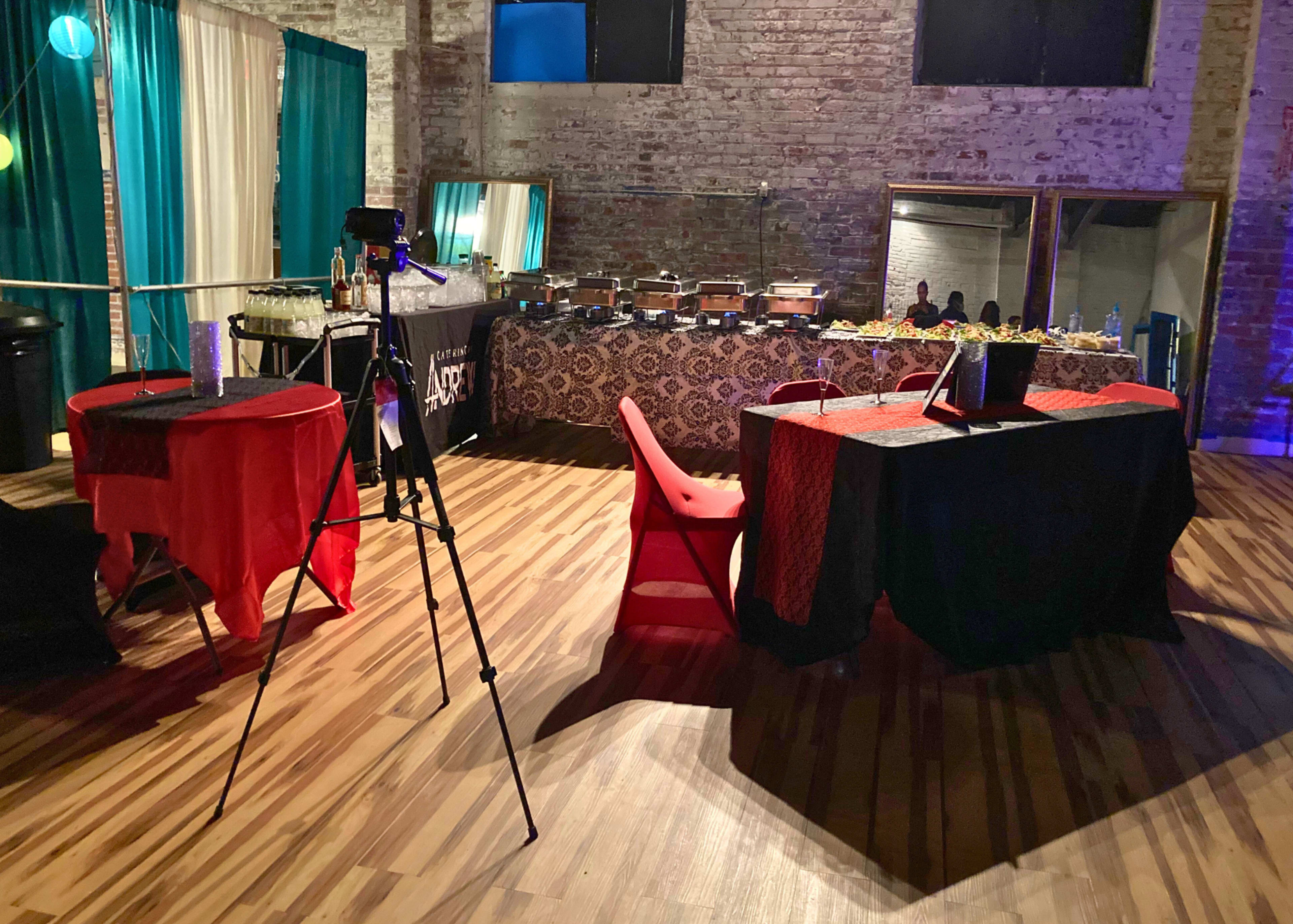 An event space with two tables, one red and one black, set up with catering dishes in the background, and a camera positioned on a tripod facing the tables.