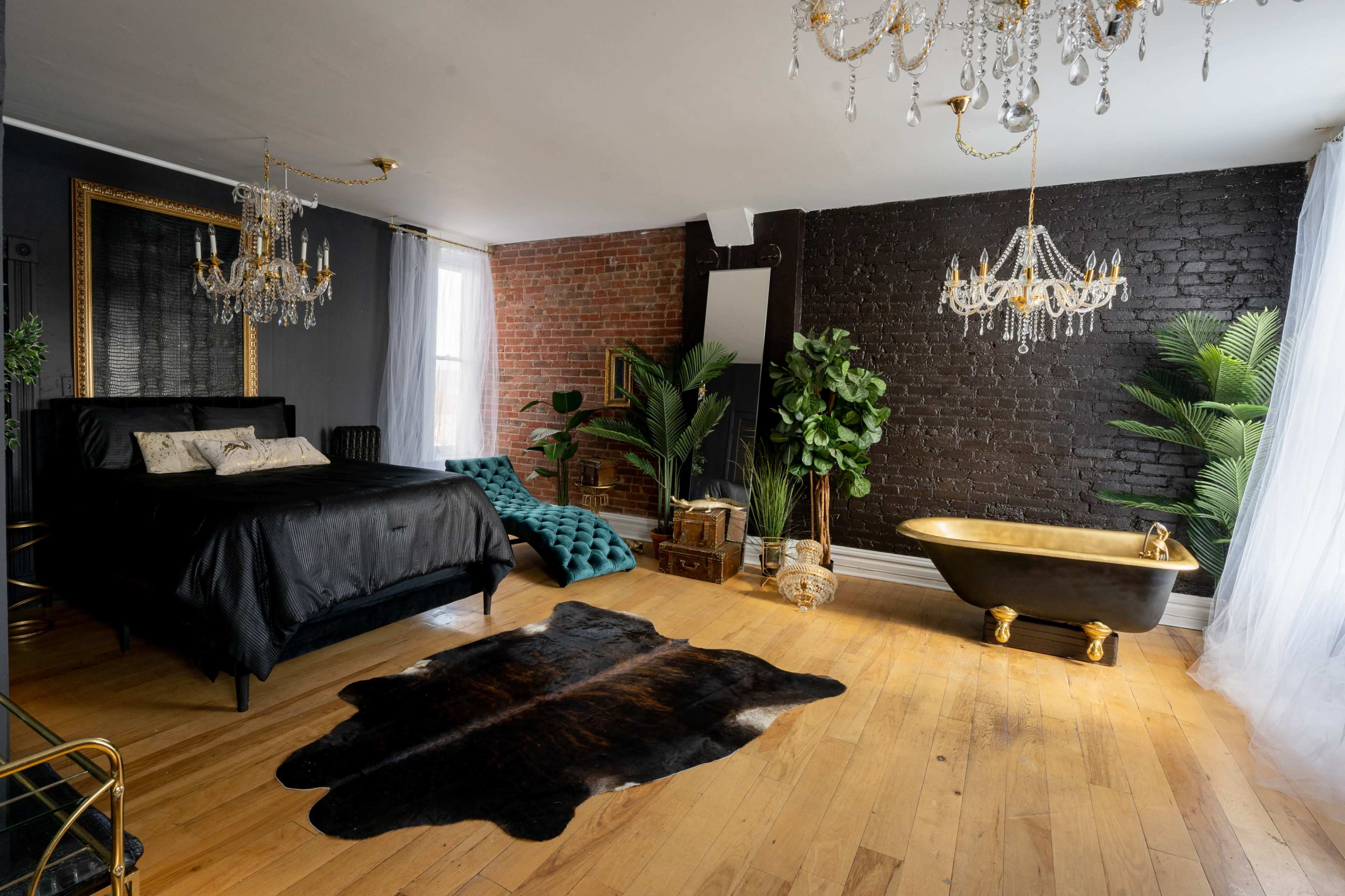 The room features a black velvet bed, a gold-accented freestanding bathtub, and a combination of exposed brick and dark walls, with greenery and decorative chandeliers enhancing the space.