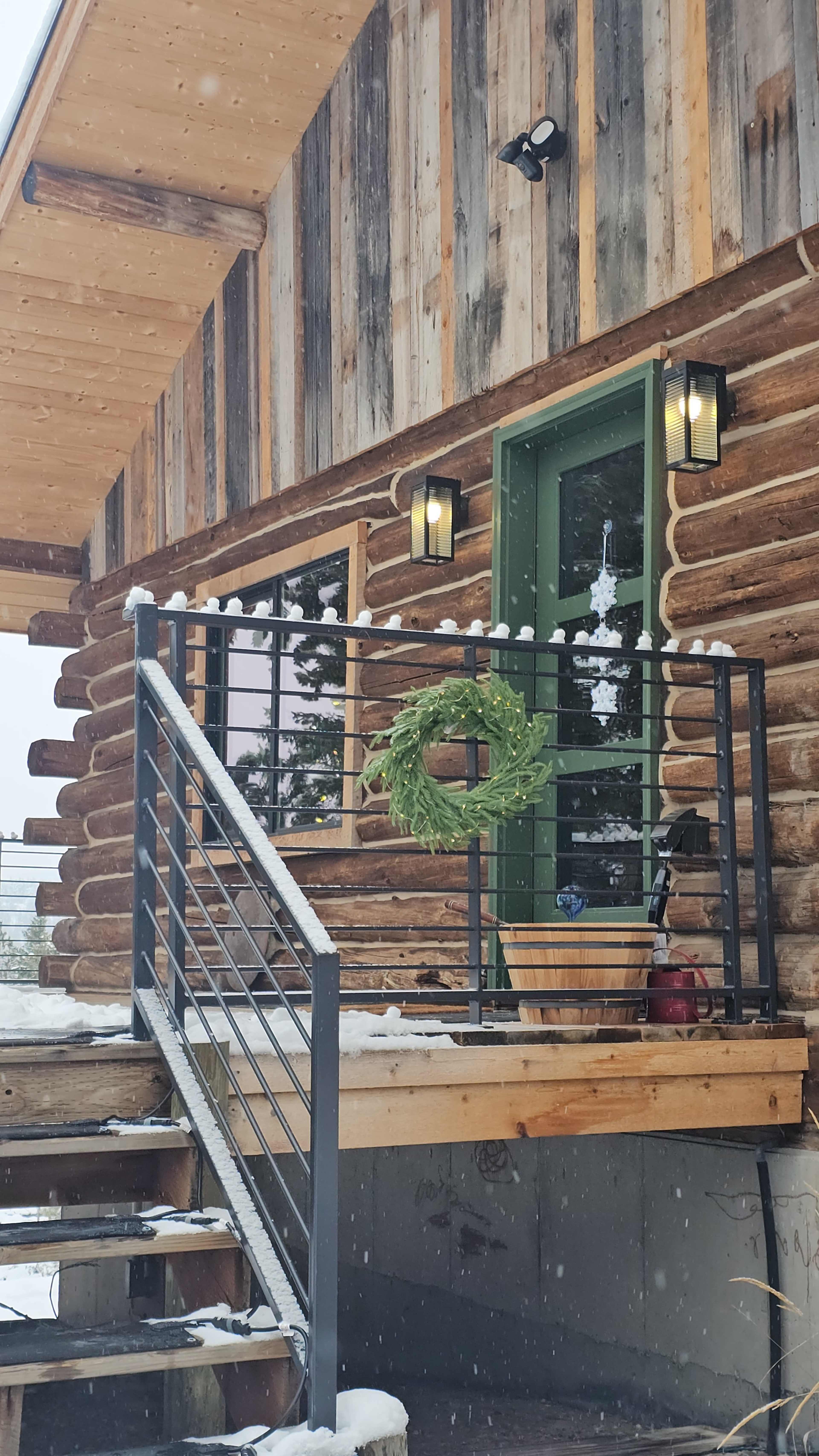The image shows a log cabin entrance with a green door, a wreath hanging on the railing, and snow falling softly on the scene.