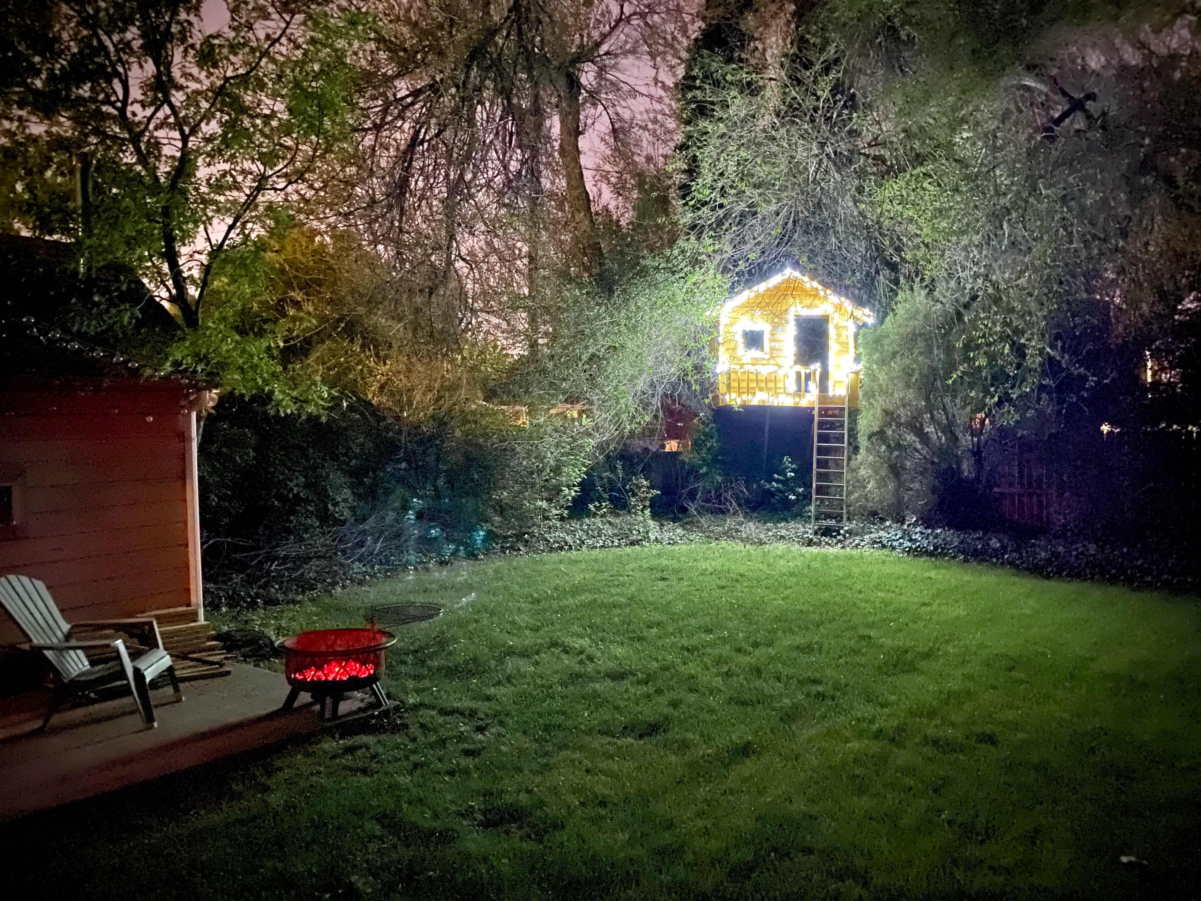 A backyard features a grassy area with a fire pit surrounded by chairs, and a brightly lit treehouse in the background.