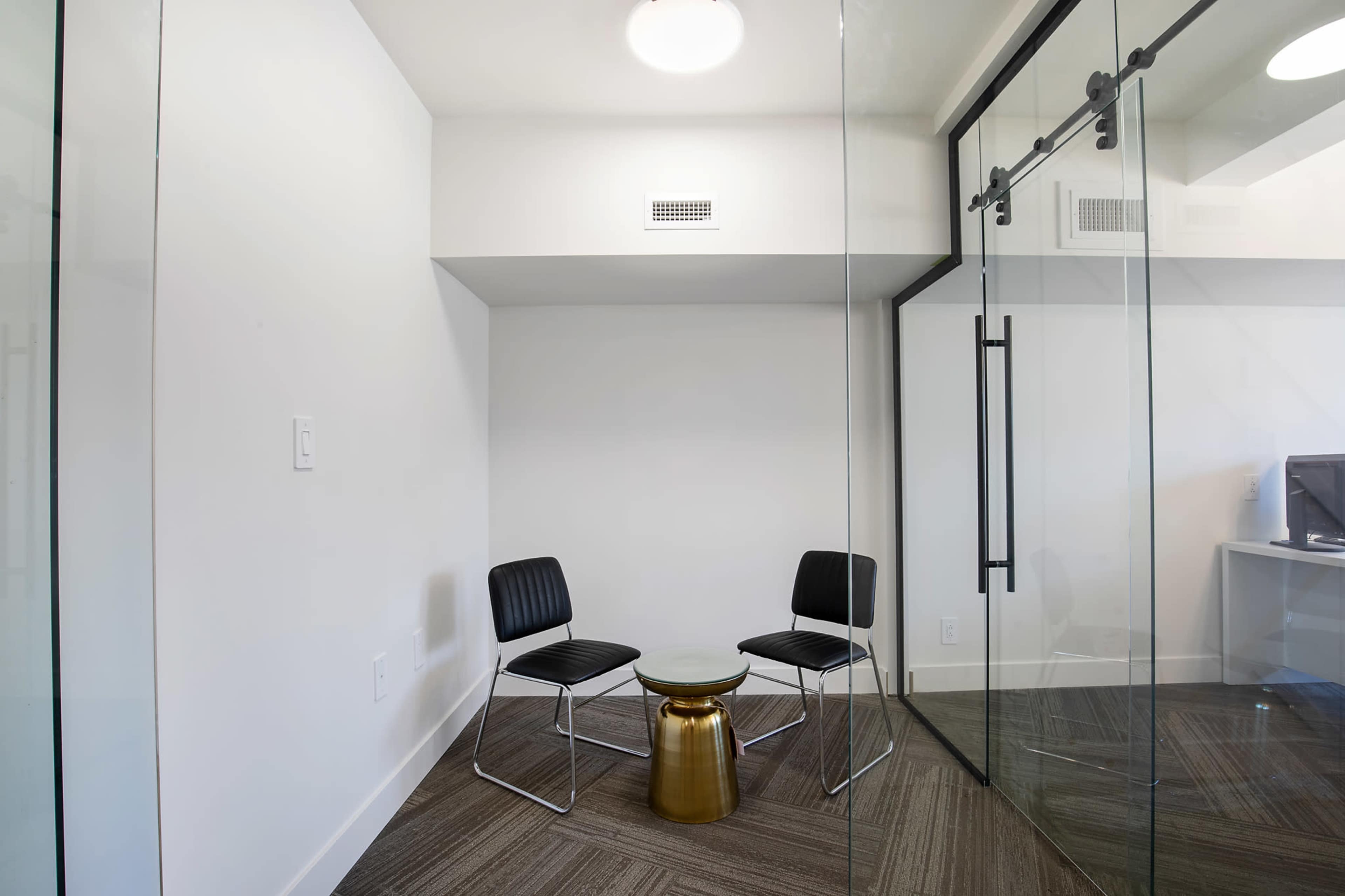 The image shows a small, glass-walled meeting room with two black chairs and a circular glass table on a carpeted floor.