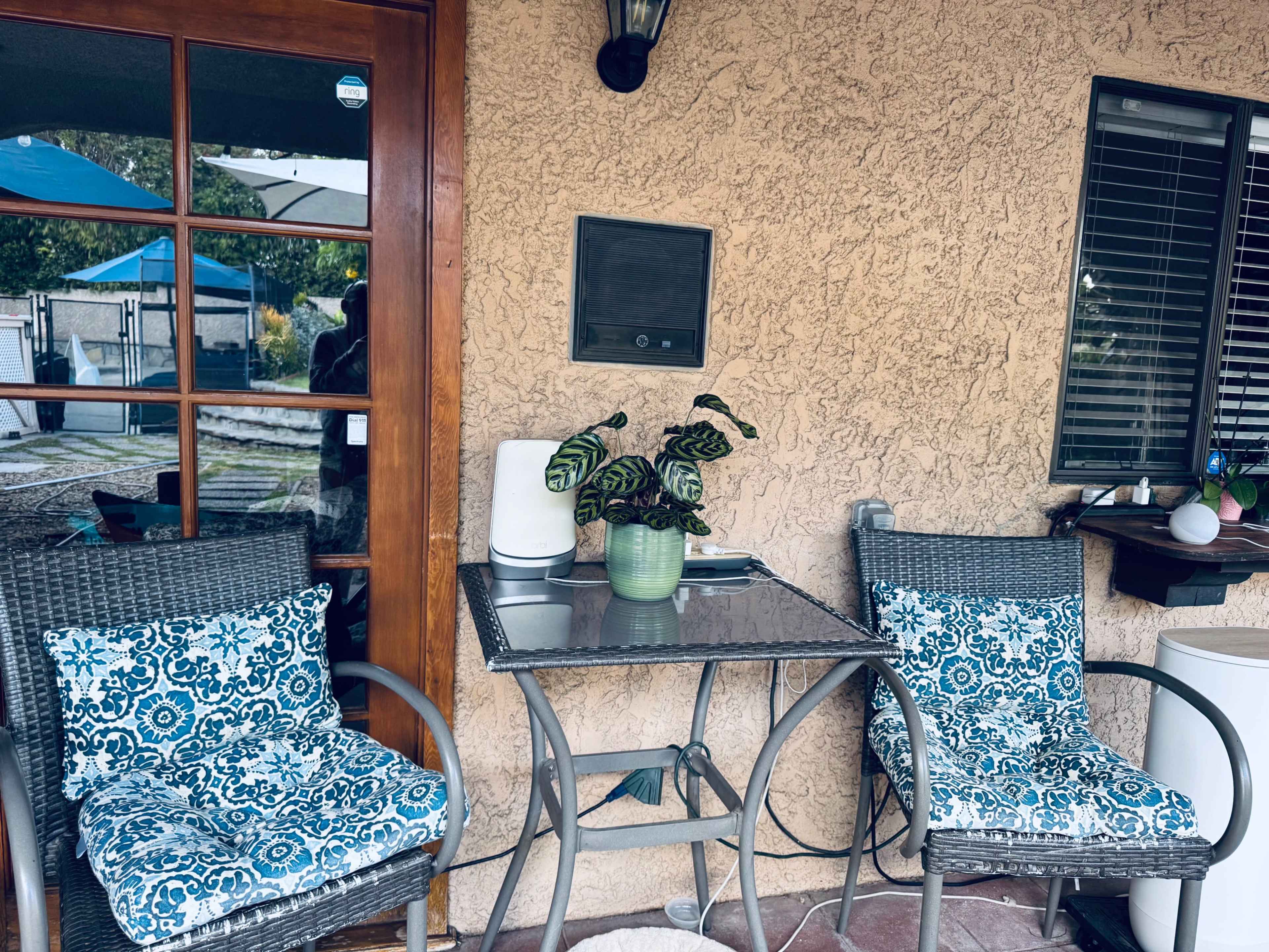 A patio area features two wicker chairs with blue patterned cushions beside a glass-top table, adorned with a potted plant.