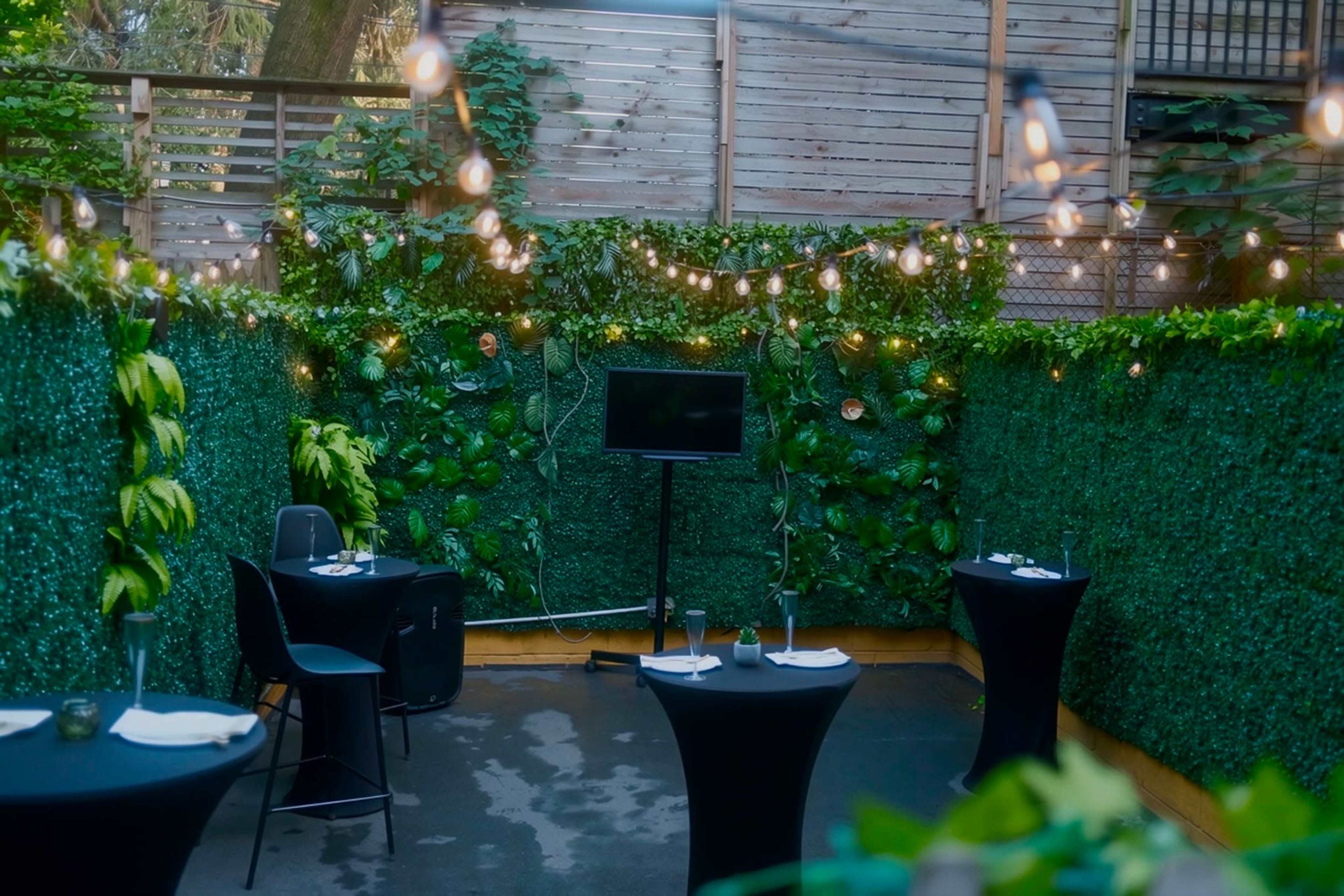 The image shows a small outdoor dining area with black tables surrounded by lush greenery and strung lights overhead.
