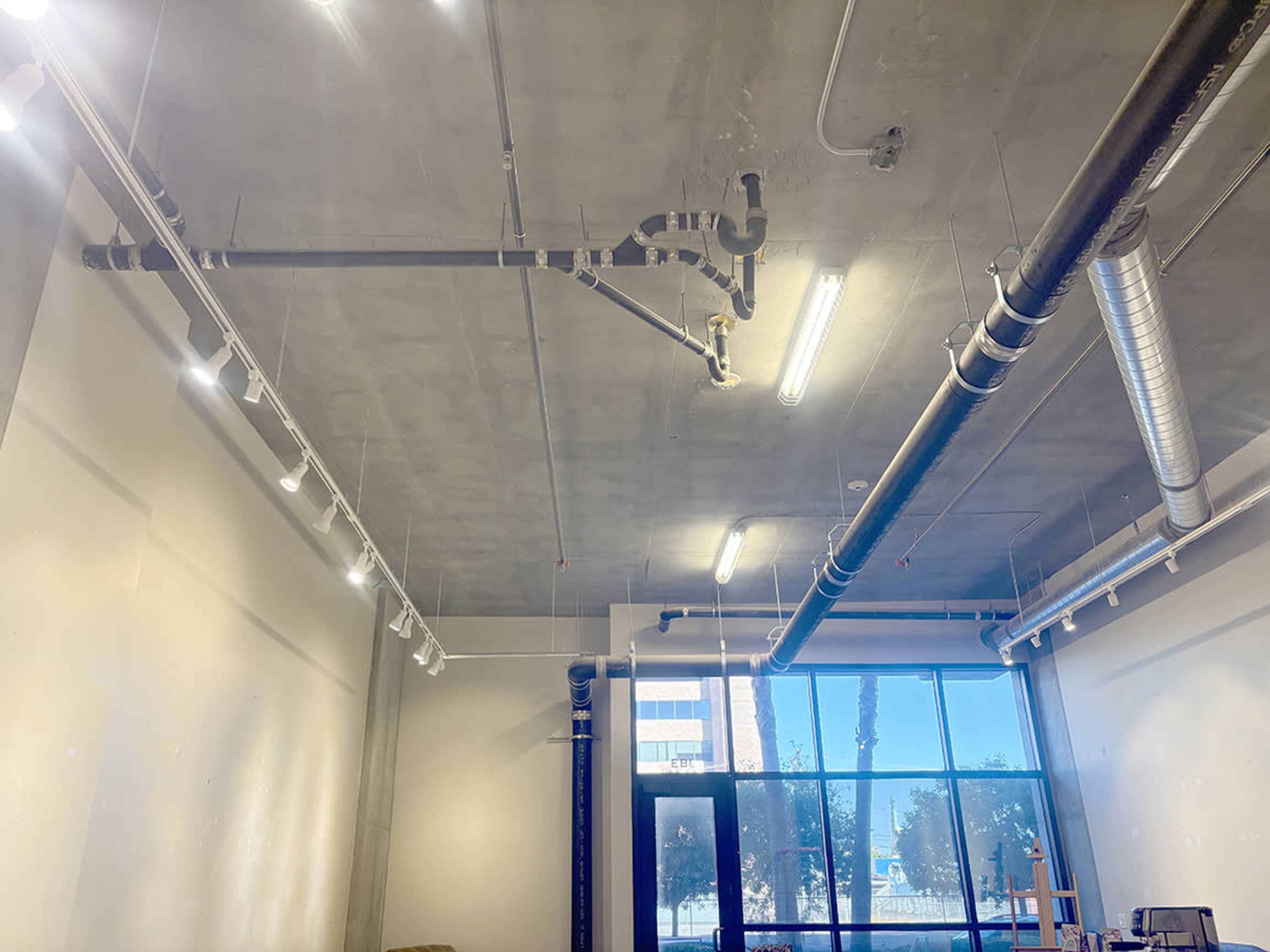 The image shows a modern interior ceiling featuring exposed pipes, ductwork, and lighting fixtures above a large window.