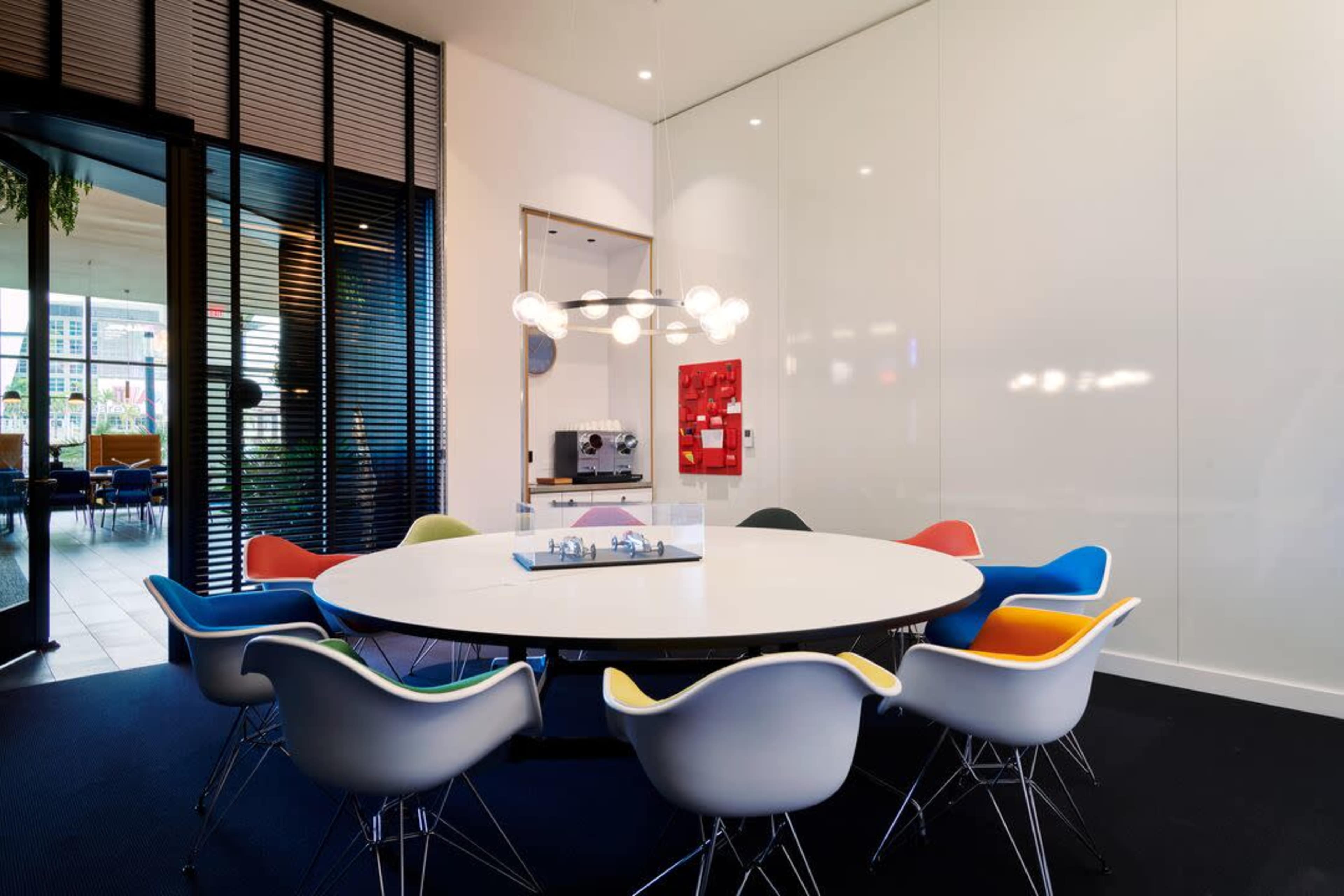 A circular conference table surrounded by colorful chairs is set in a modern meeting room with large windows and minimalistic decor.