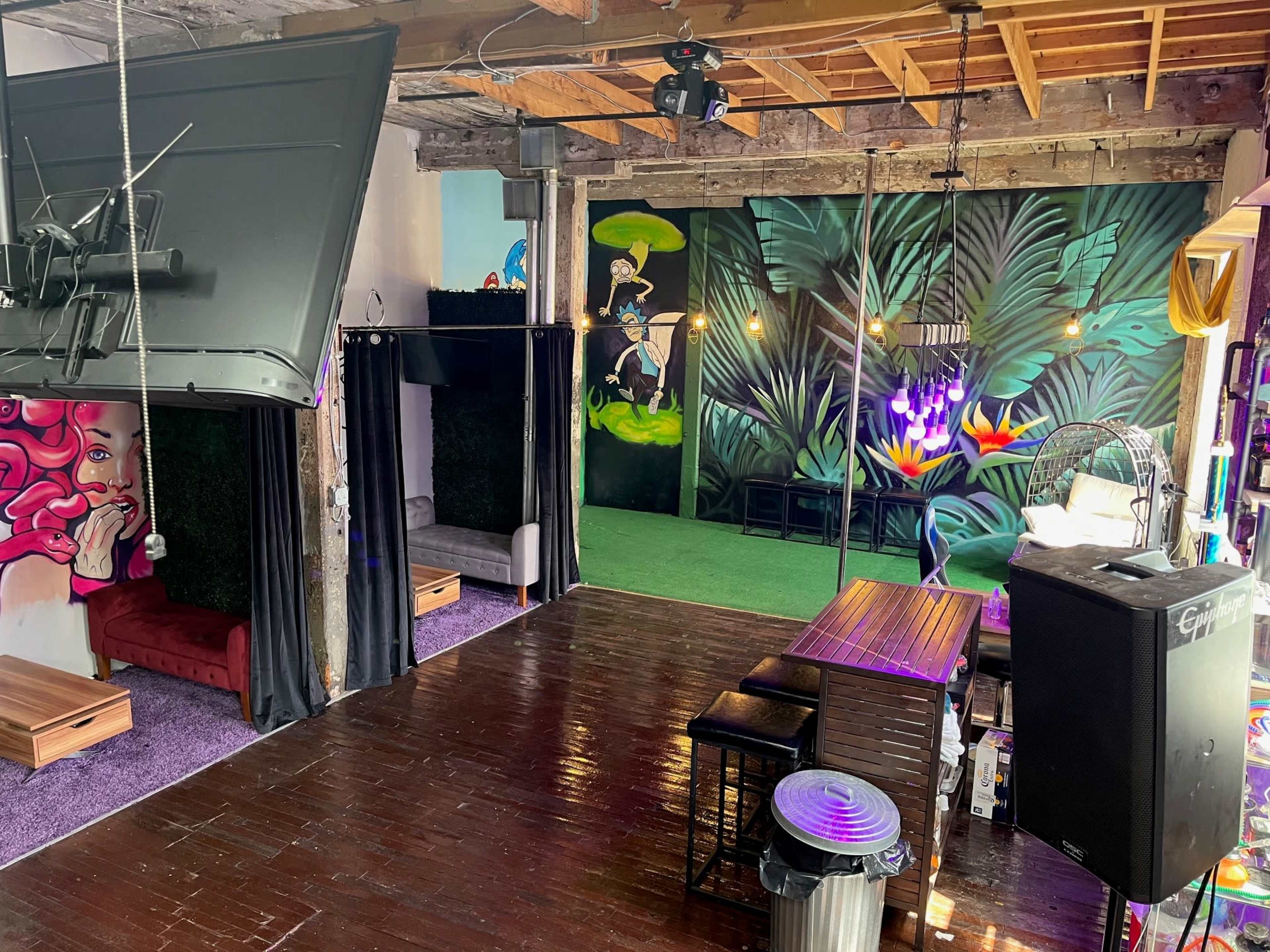 An indoor space with colorful murals, a wooden floor, seating areas, and a lush green backdrop.