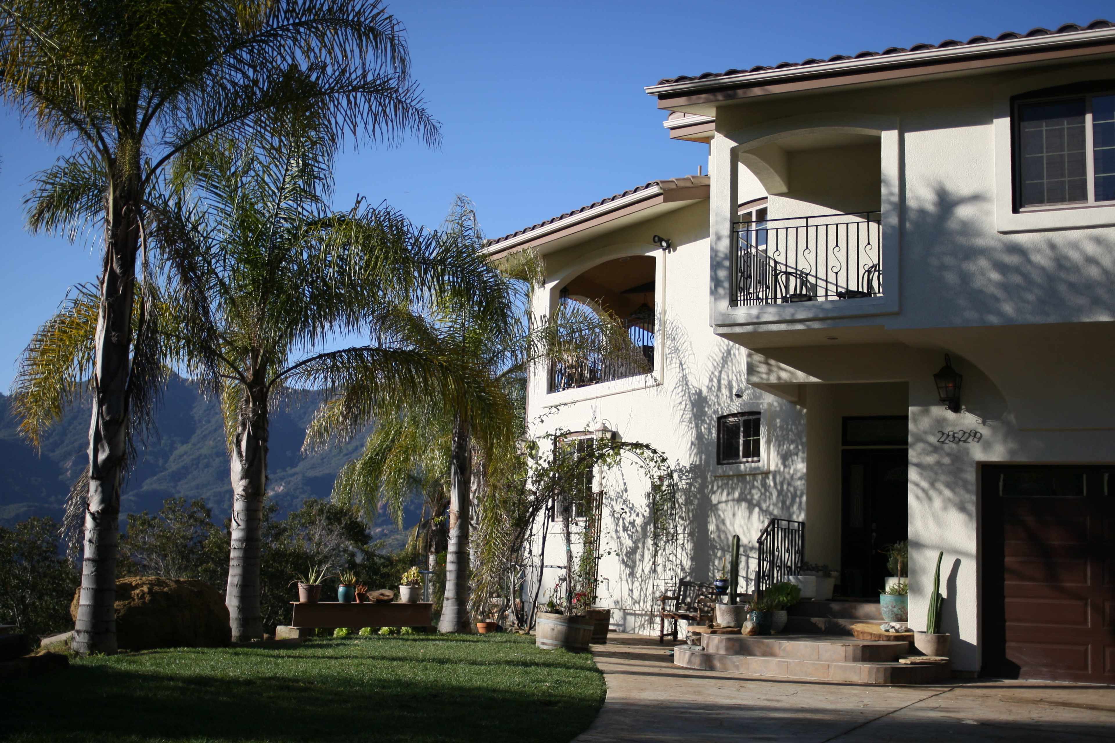 A two-story house with a balcony overlooks a landscaped yard featuring palm trees and mountain views in the background.