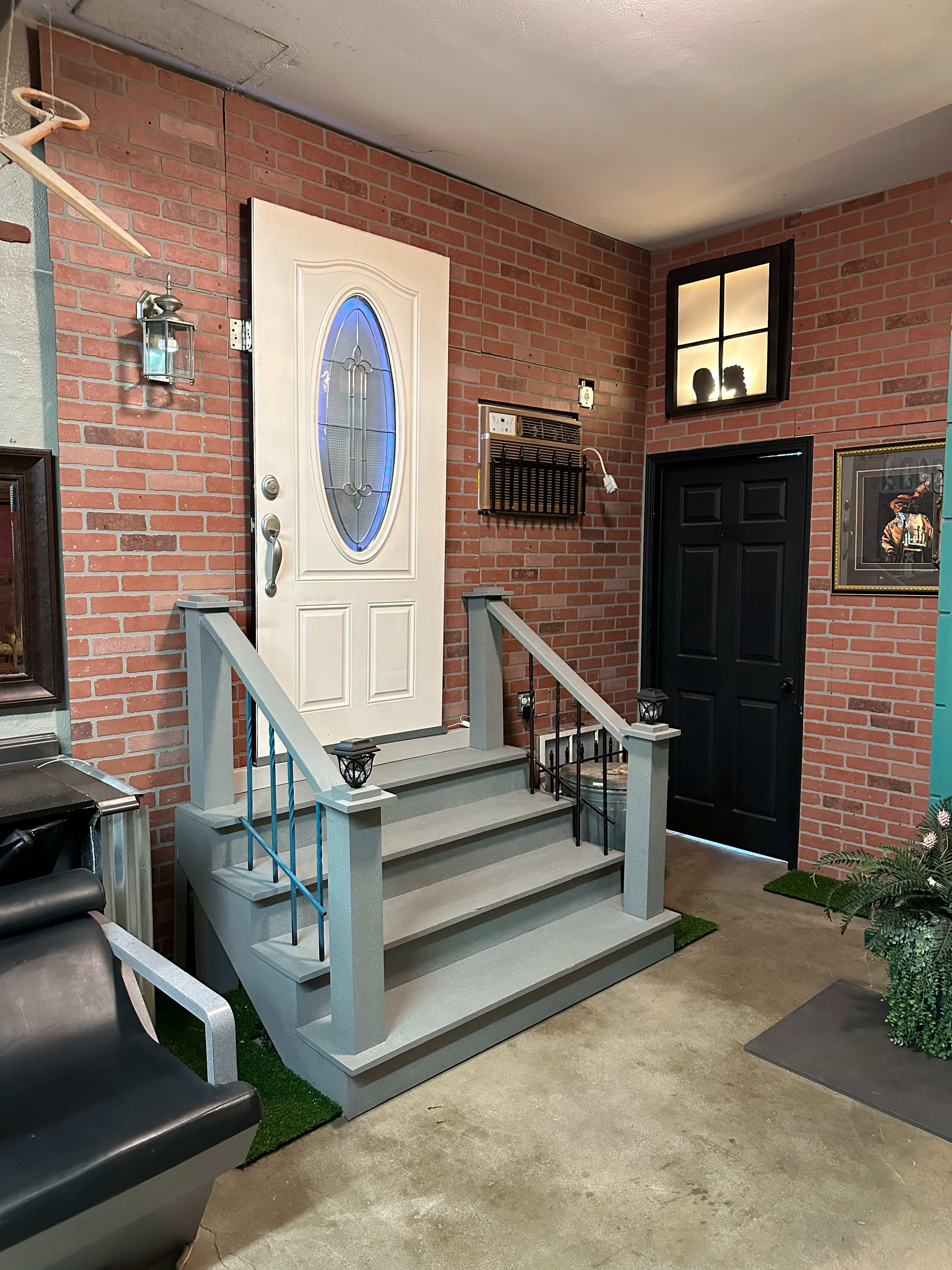 A small entryway with a white door, a set of gray stairs, and brick walls, featuring a decorative light and a black door on the side.