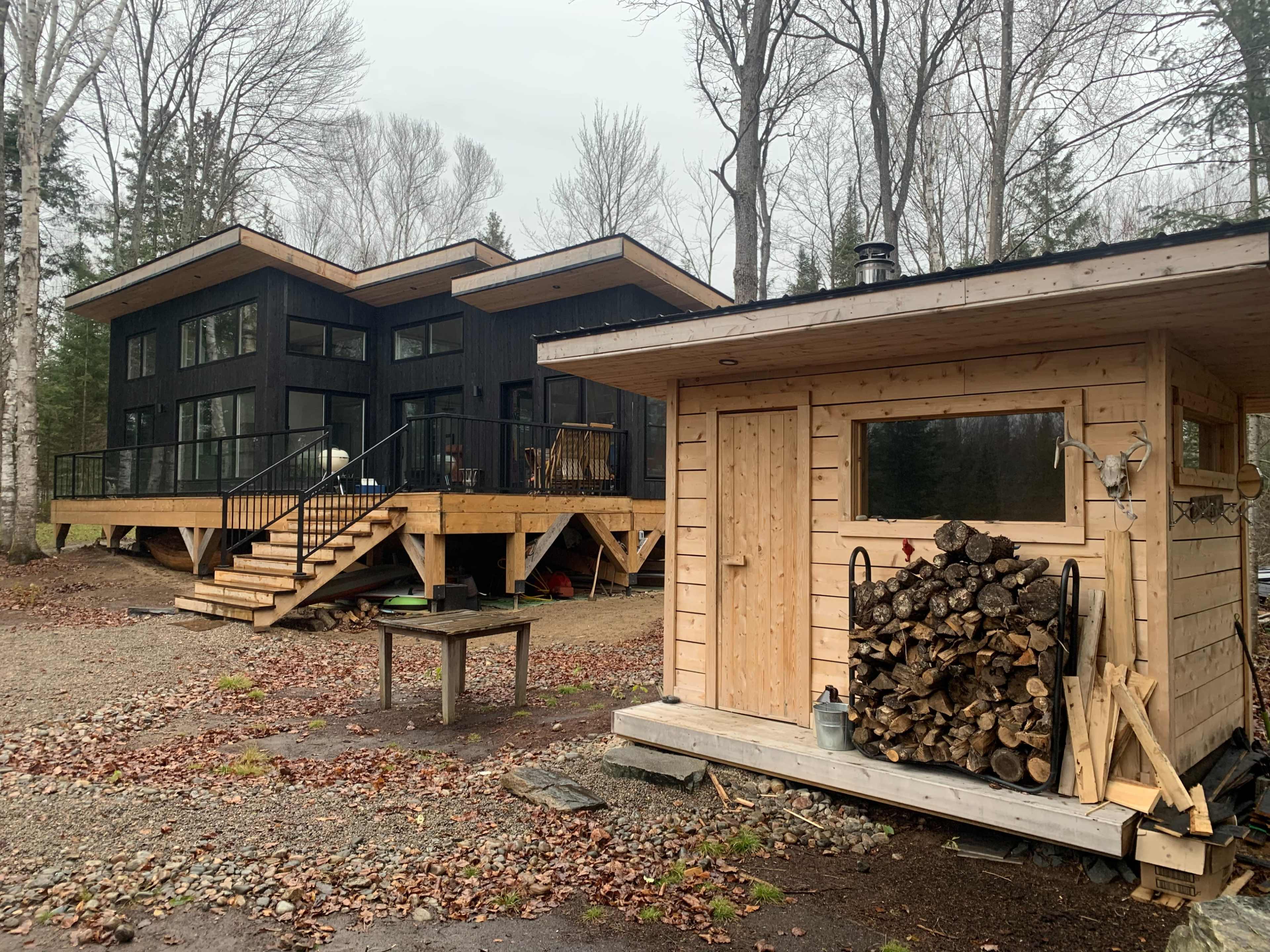 A modern, dark wooden house with large windows sits on a raised deck beside a smaller wooden shed stacked with firewood, surrounded by trees in a gravel clearing.