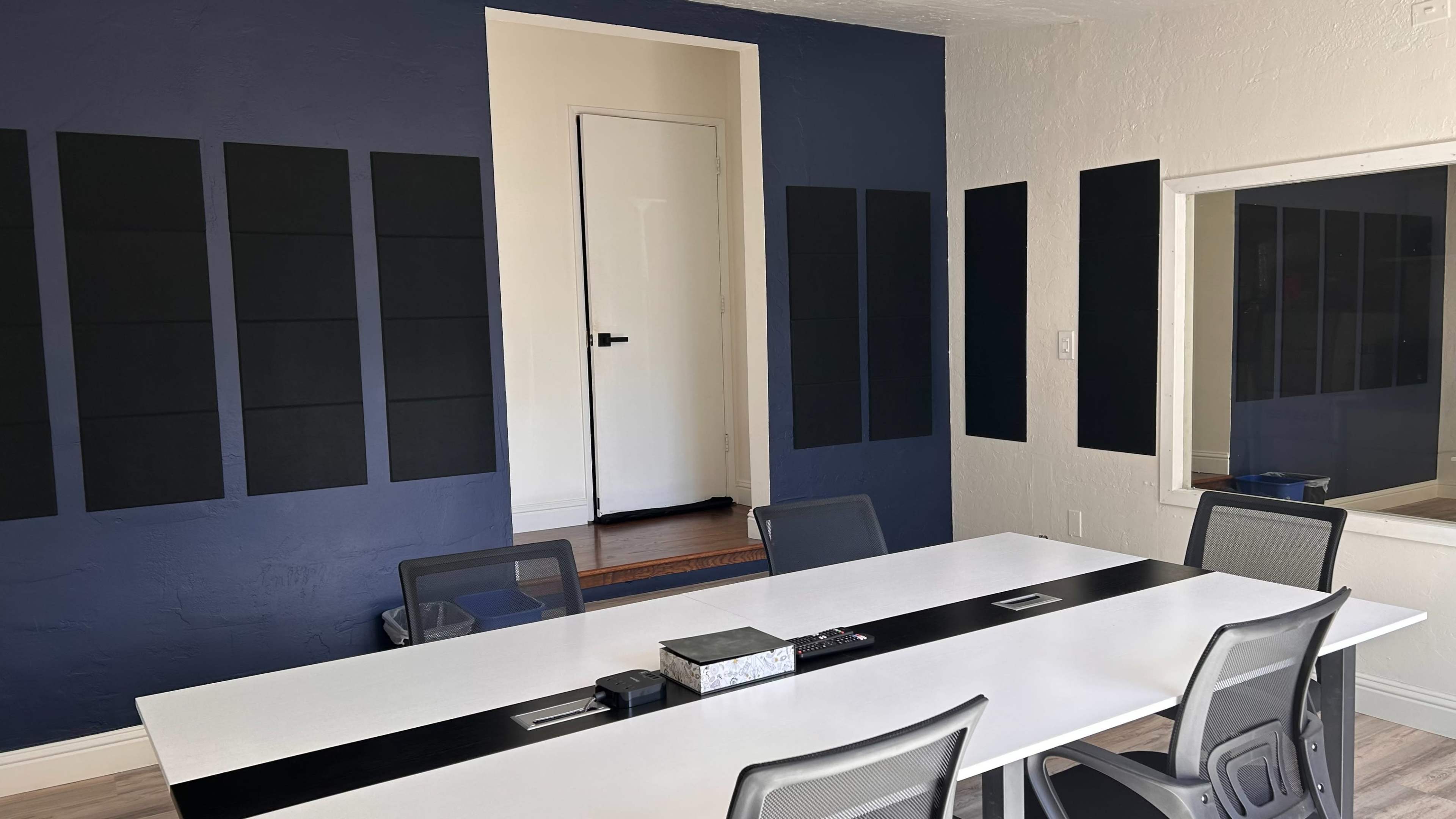 A conference room with a long white table and gray chairs, featuring dark wall panels and an open door leading to another room.