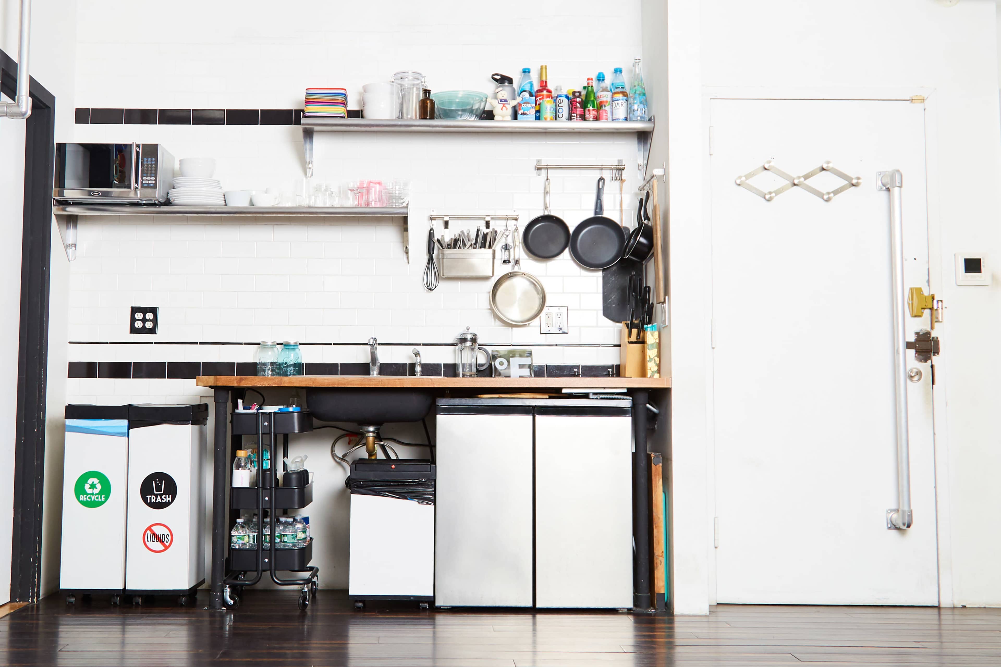 The image shows a compact kitchen area with a sink, microwave, shelving with various kitchen items, and an entrance door.