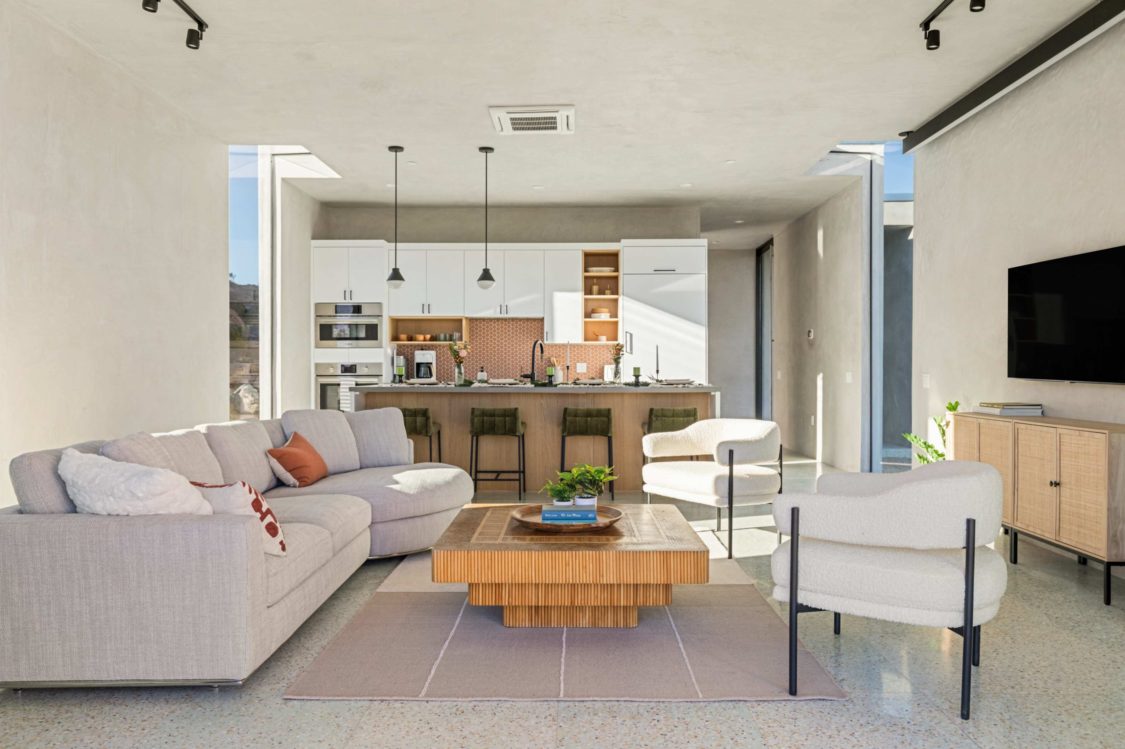 A modern living room features a sectional sofa, a wooden coffee table, and a kitchen area with white cabinetry and bar stools in the background.