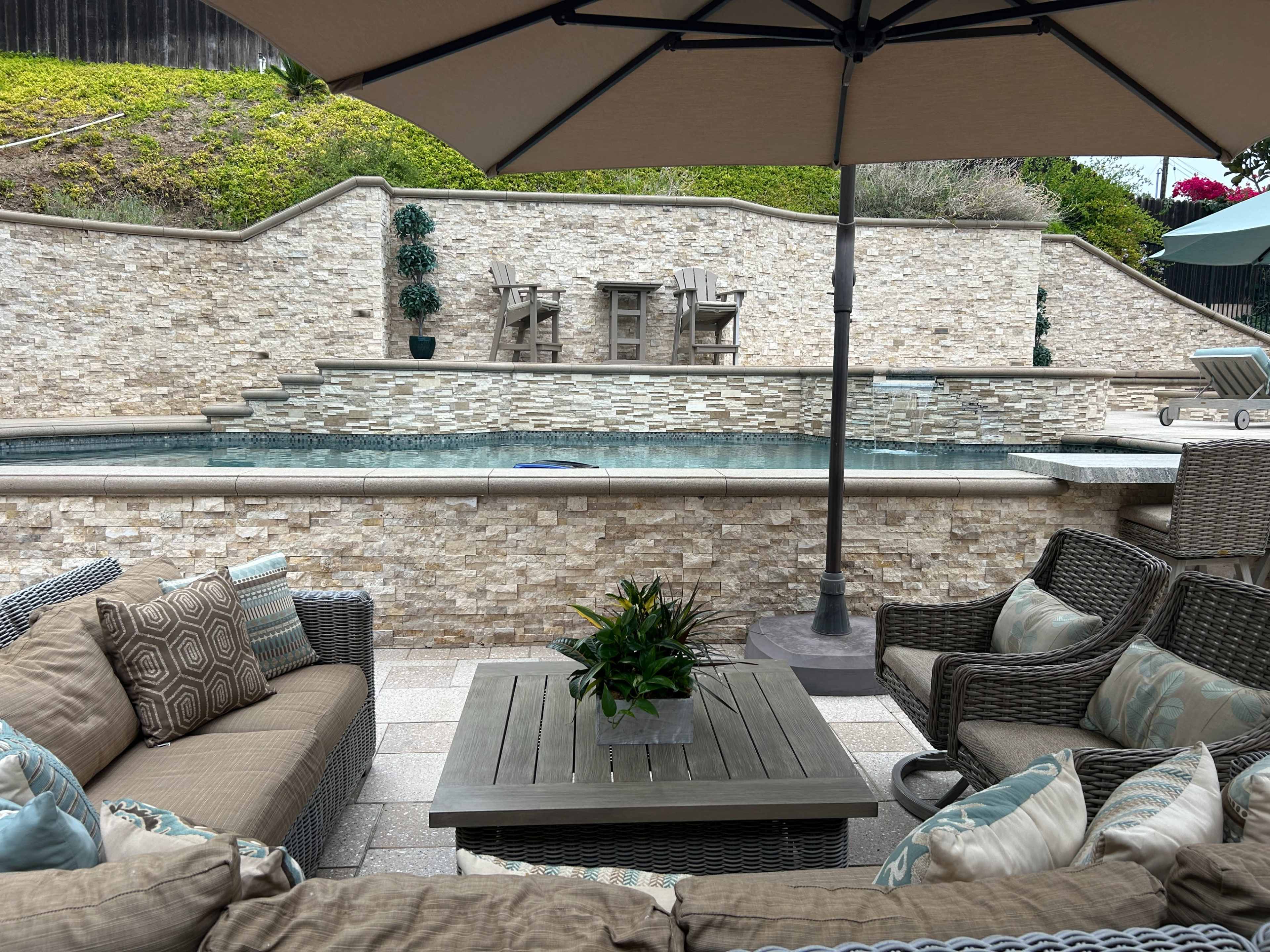 The image shows a patio area with a section of a swimming pool, surrounded by stone walls and furnished with outdoor seating and a dining table under an umbrella.