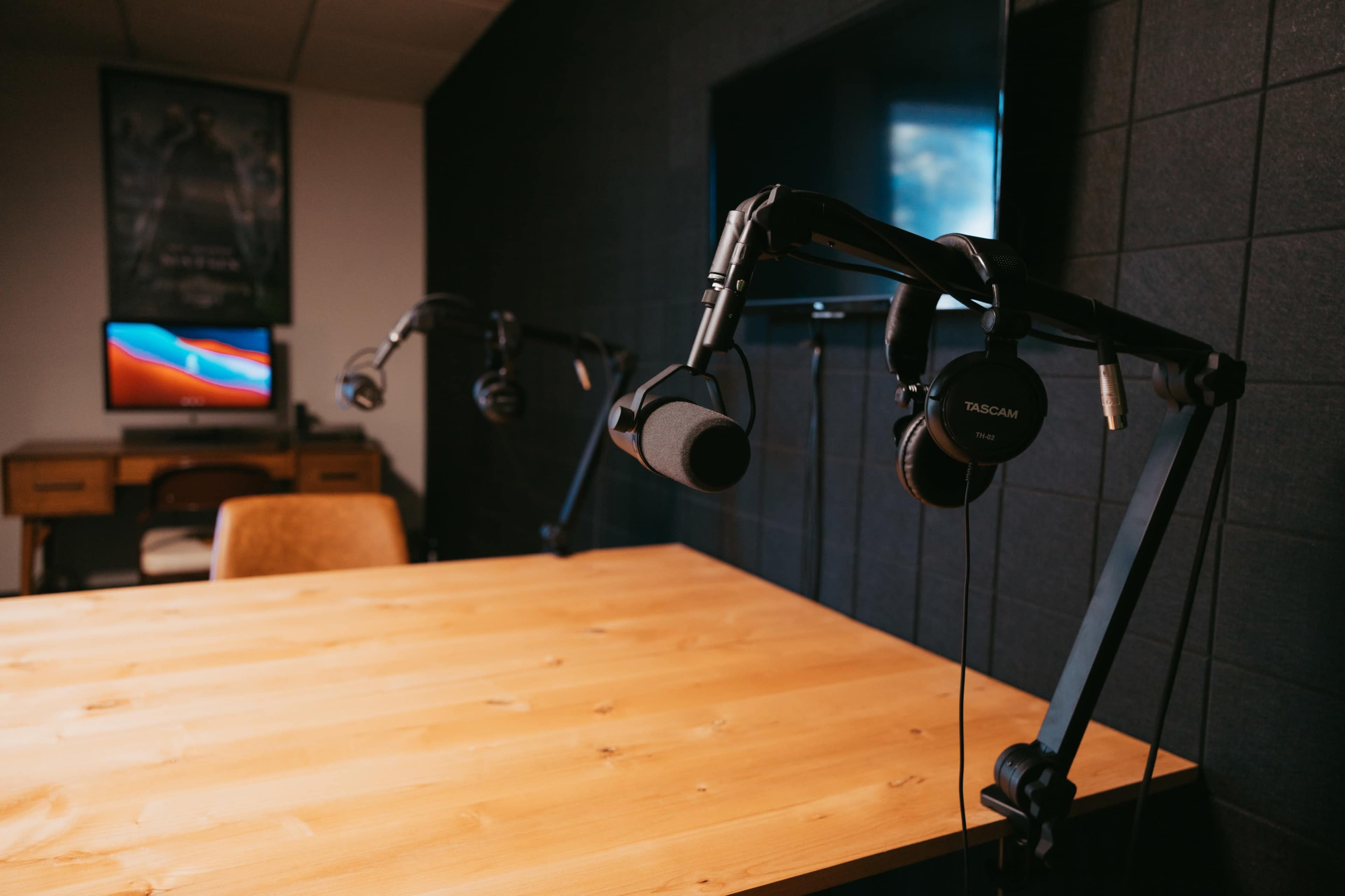 A recording studio setup with a wooden table, microphones mounted on adjustable arms, and a computer in the background.