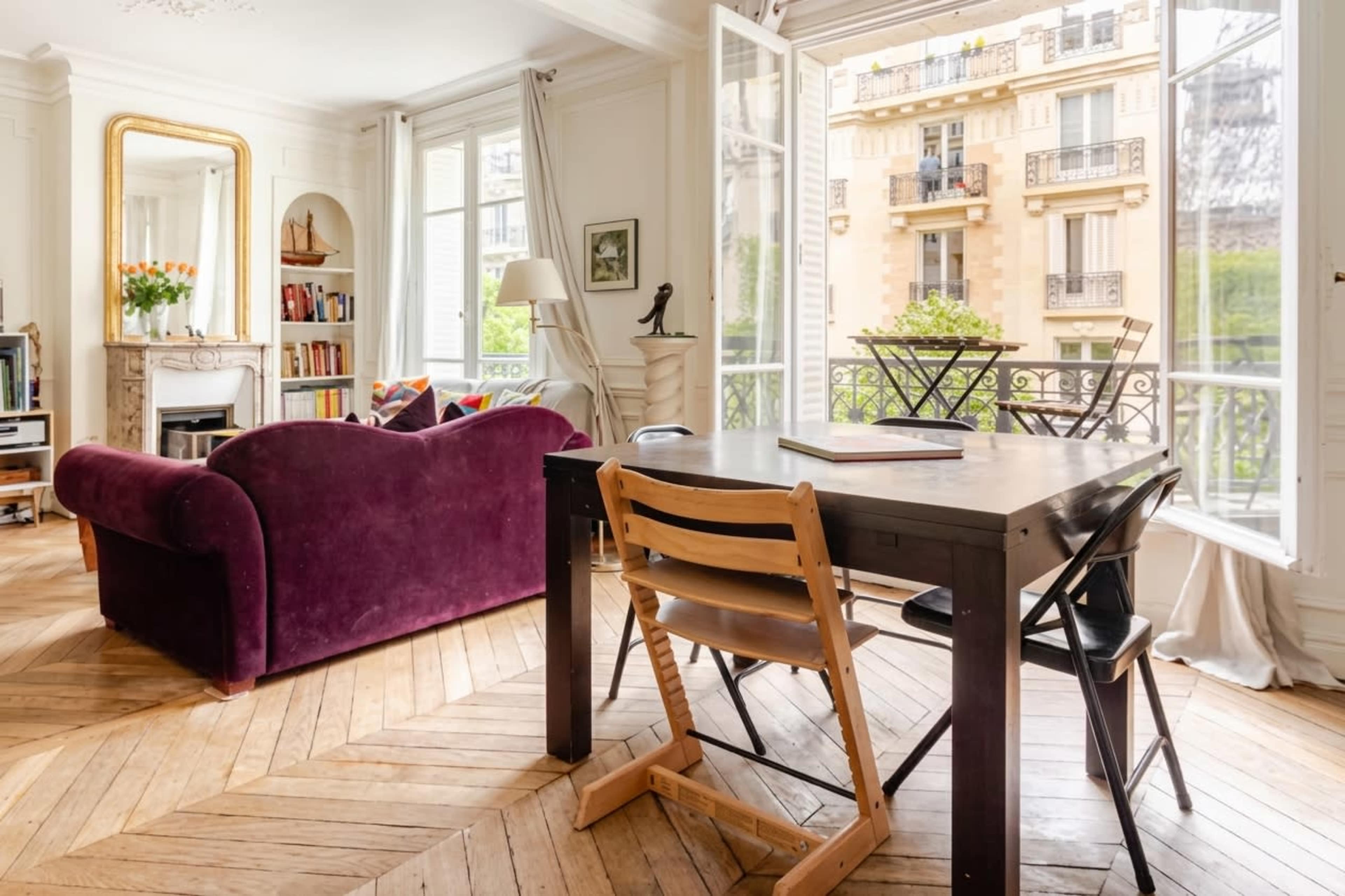 A cozy apartment features a purple couch, a wooden dining table with chairs, and large windows open to a balcony view.
