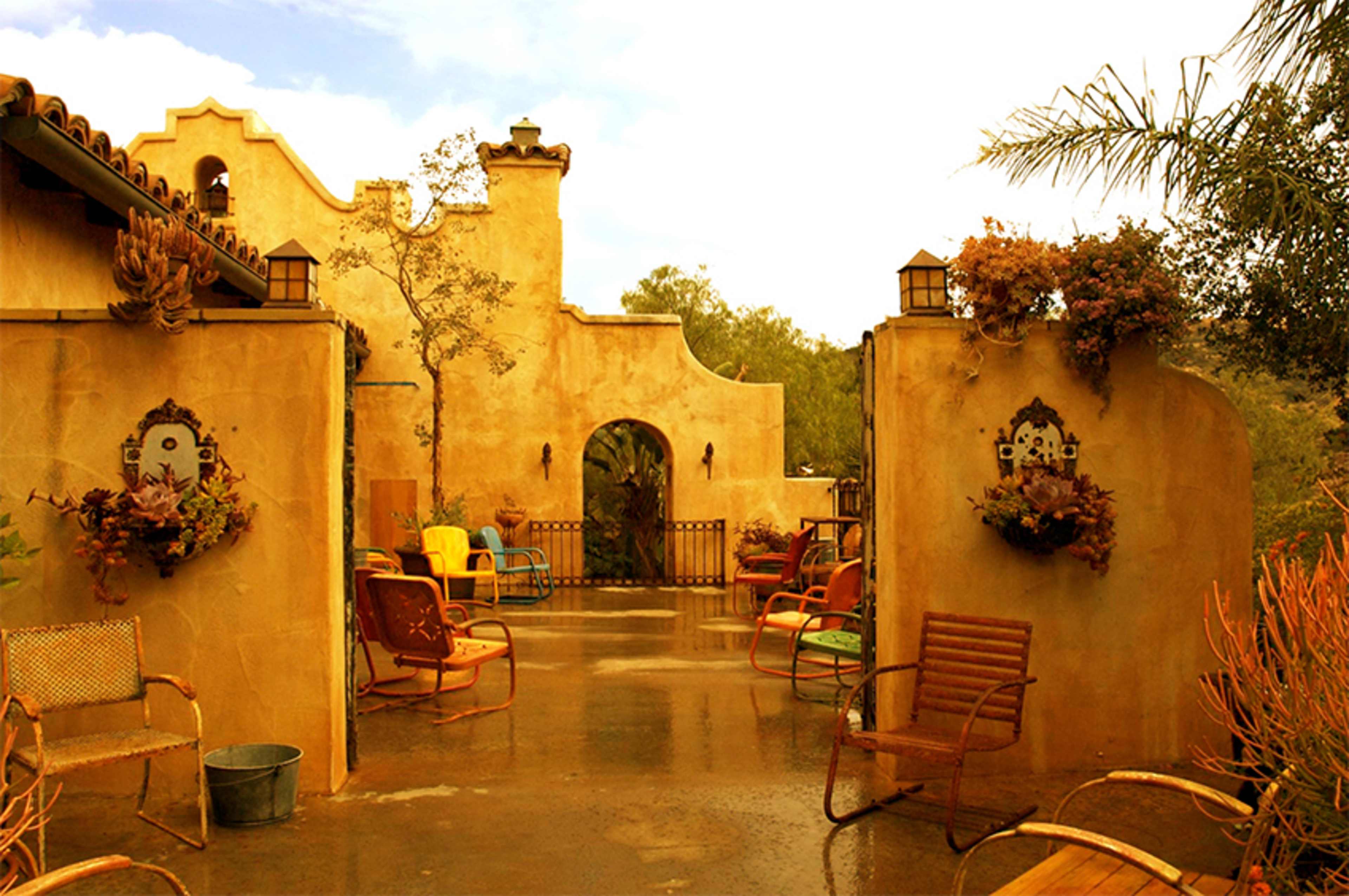 The scene features a courtyard with colorful chairs and potted plants set against a textured, earth-toned wall and an arched entrance.