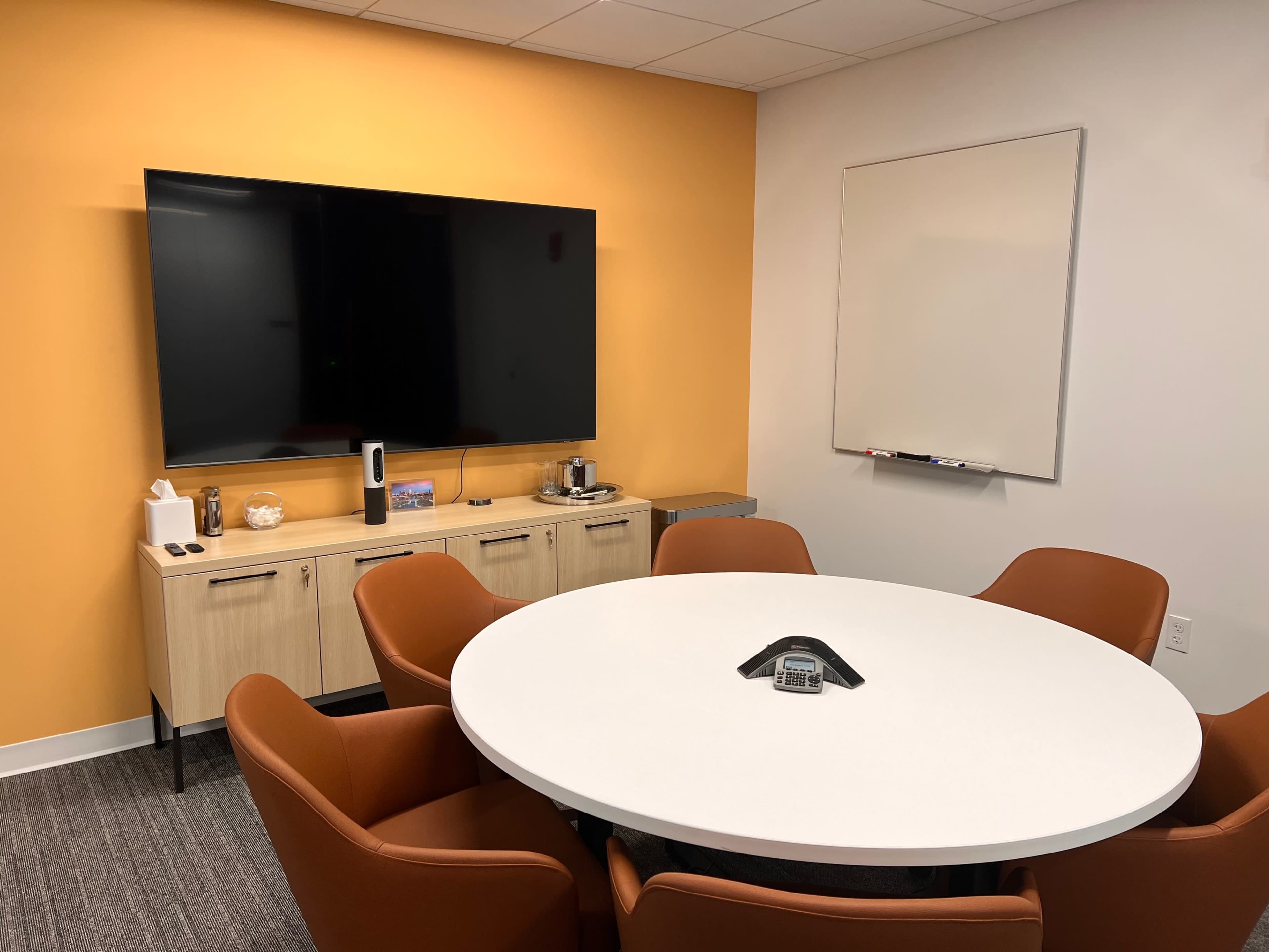 A modern conference room featuring a round white table surrounded by orange chairs, a large wall-mounted television, and an empty whiteboard.