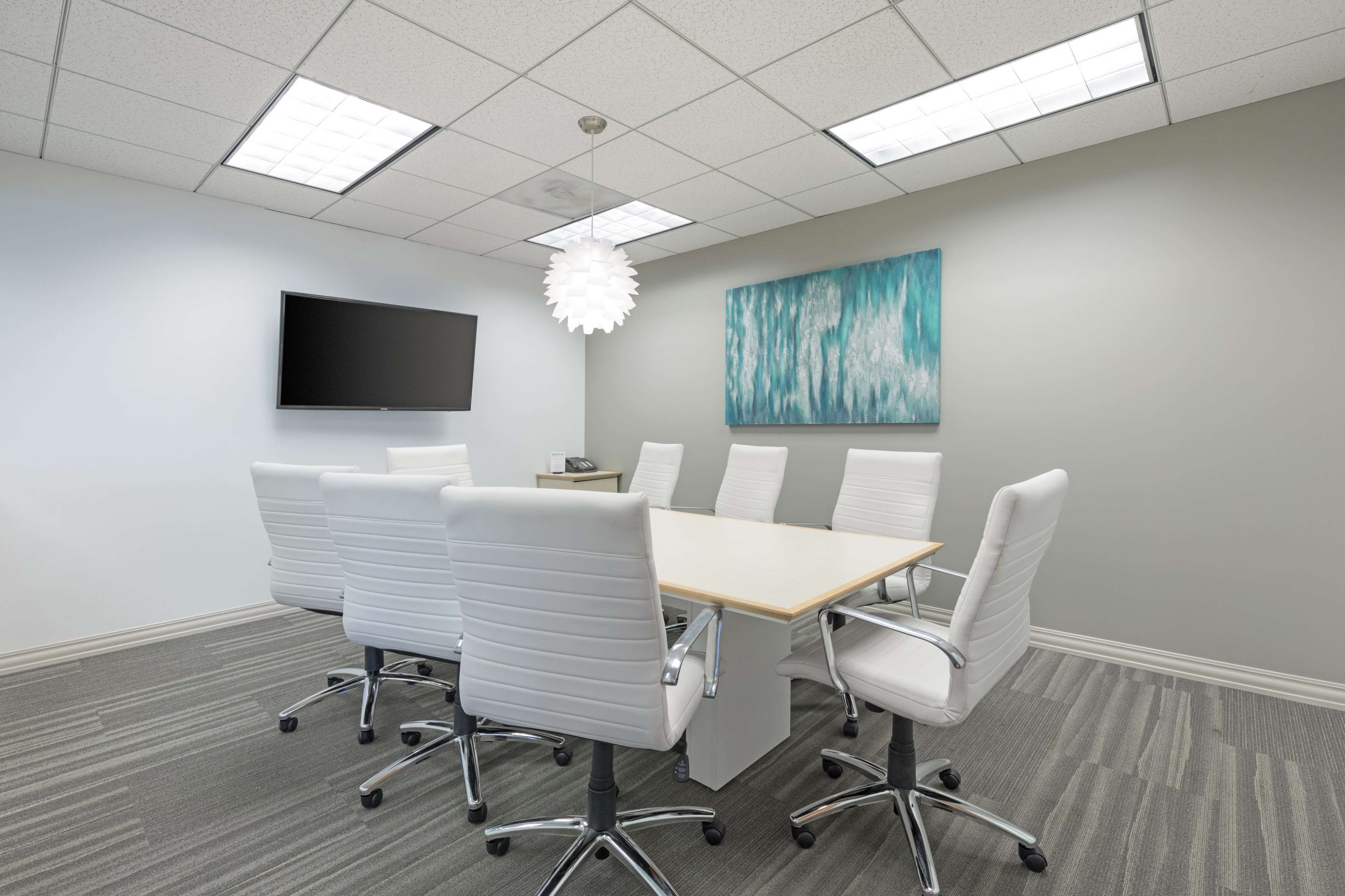 A conference room features a large rectangular table surrounded by six white chairs, with a wall-mounted television and a large blue abstract painting.