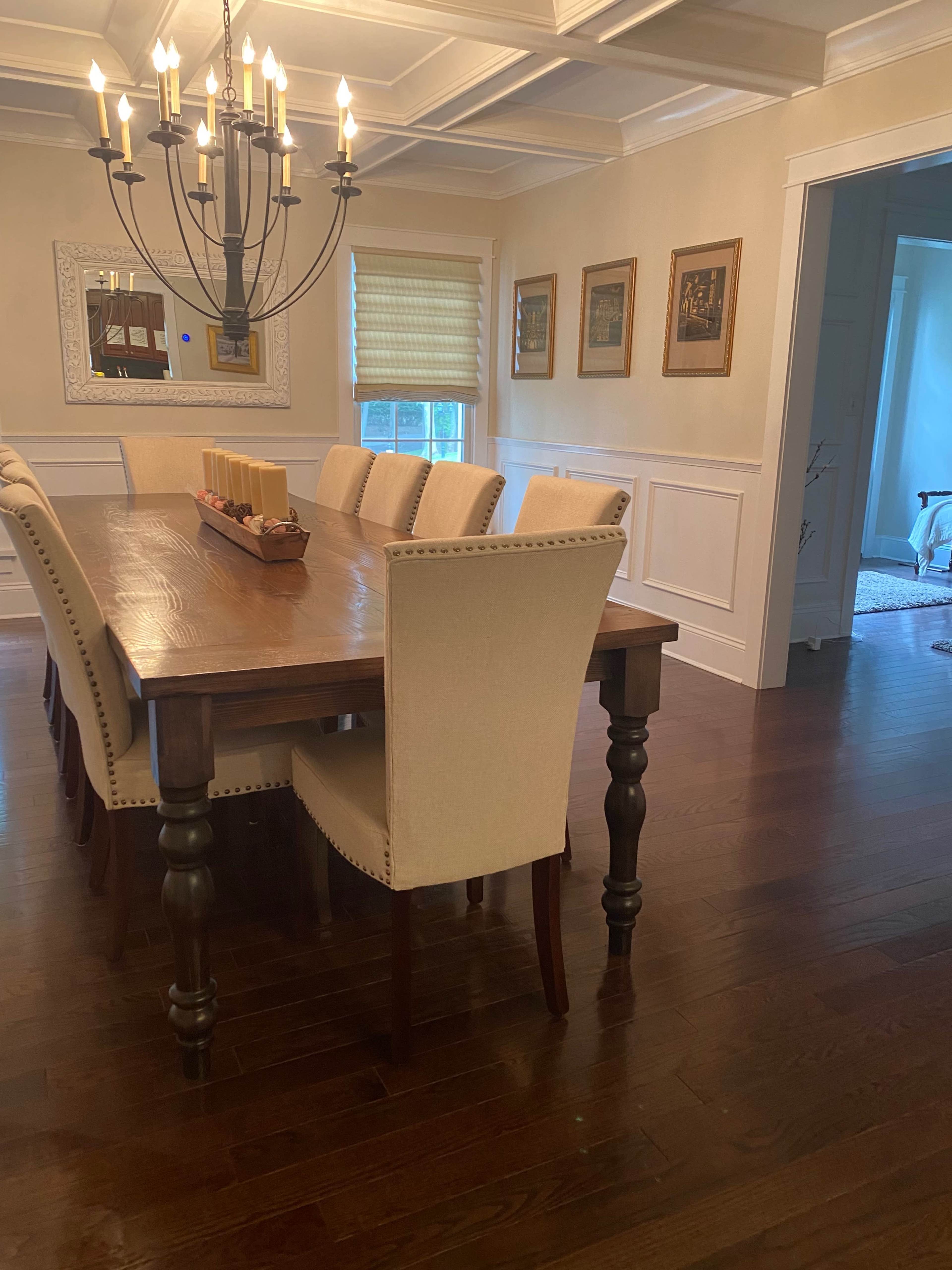 A large wooden dining table with beige upholstered chairs is positioned under a chandelier in a well-lit dining room.