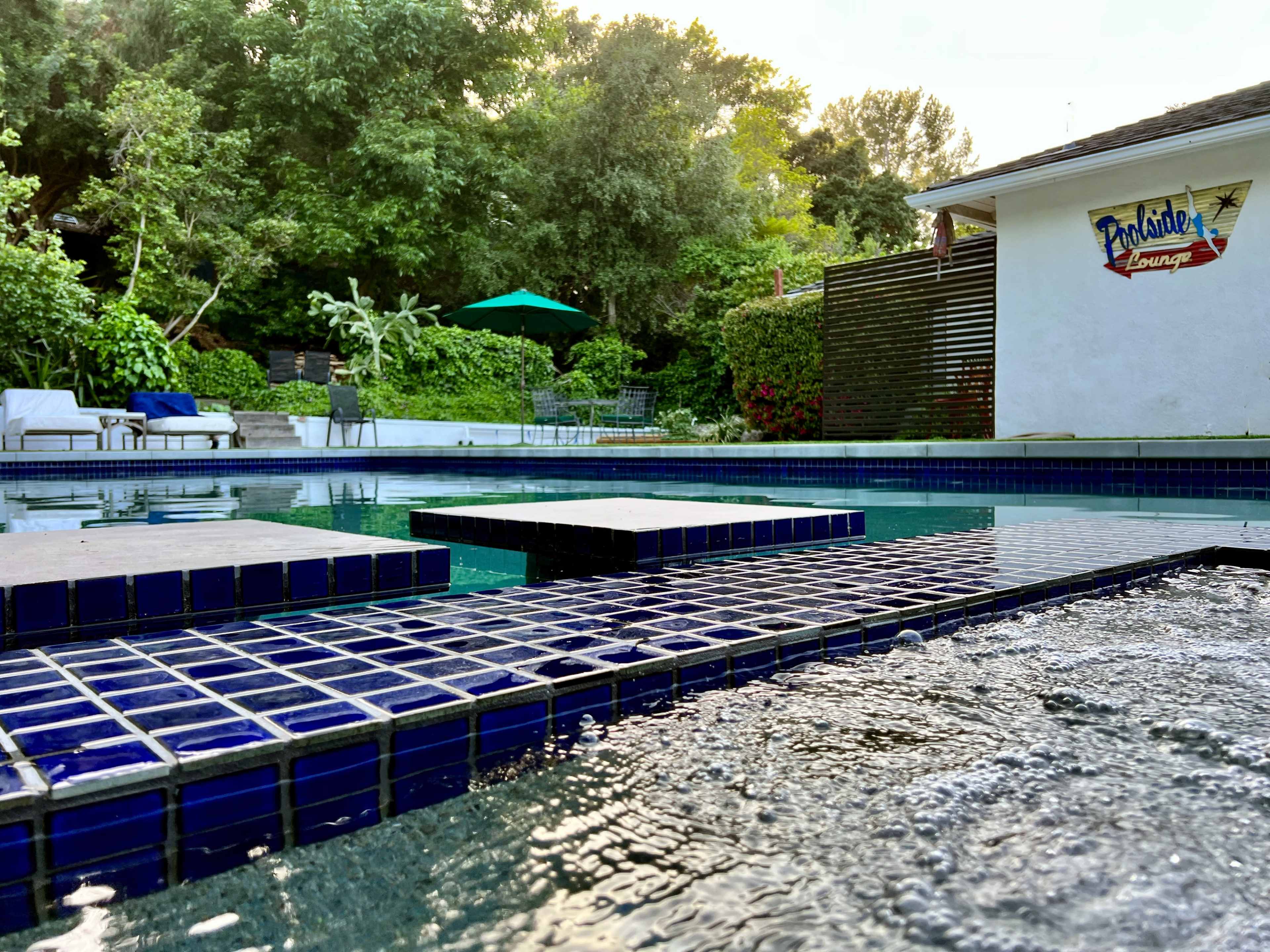 A swimming pool with dark blue tile borders and stepping stones, surrounded by lush greenery and a nearby lounge area.