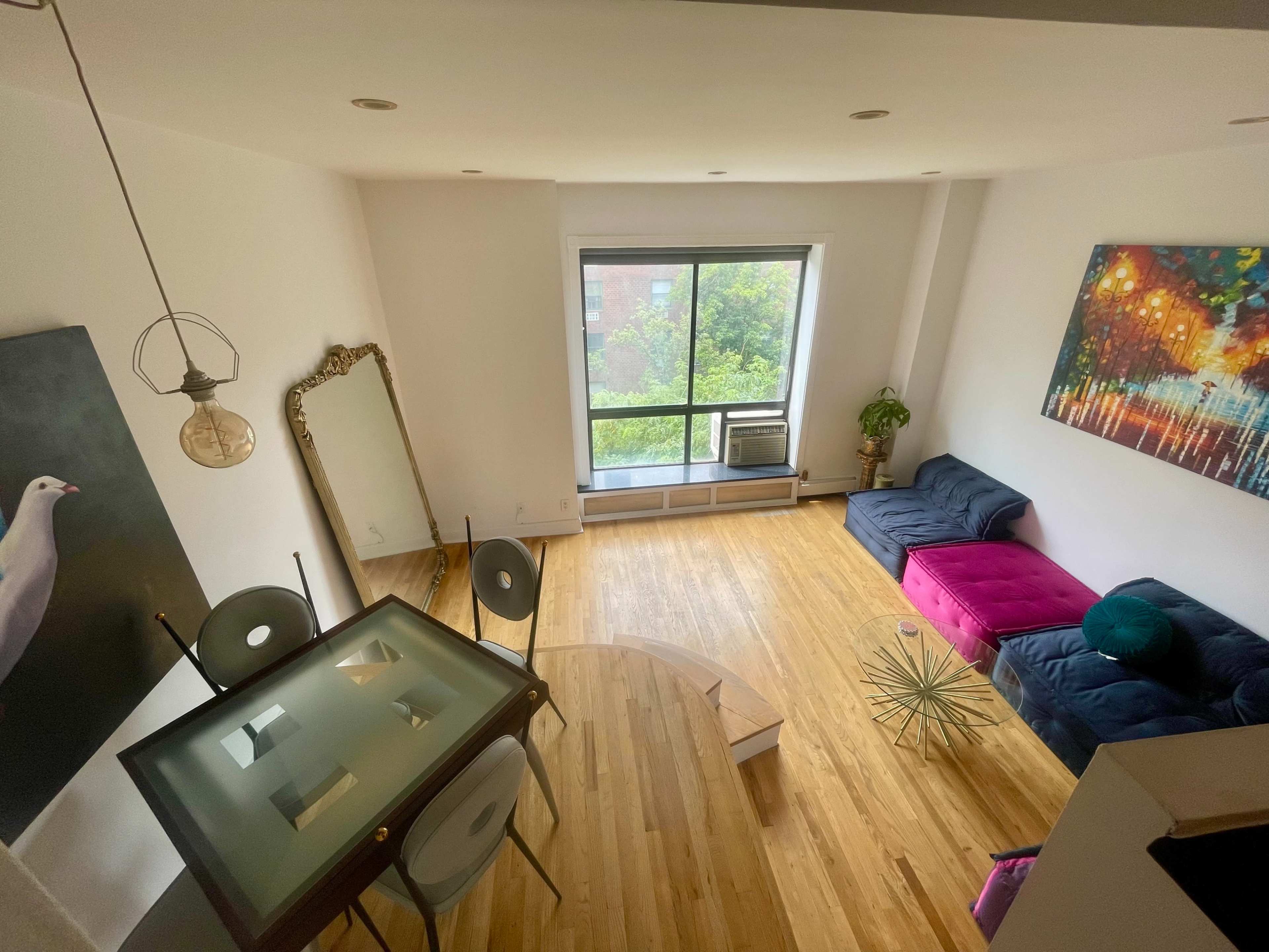 A spacious, well-lit room features a large window, a dark blue couch with a pink section, a round wooden coffee table, and a dining table with transparent chairs.