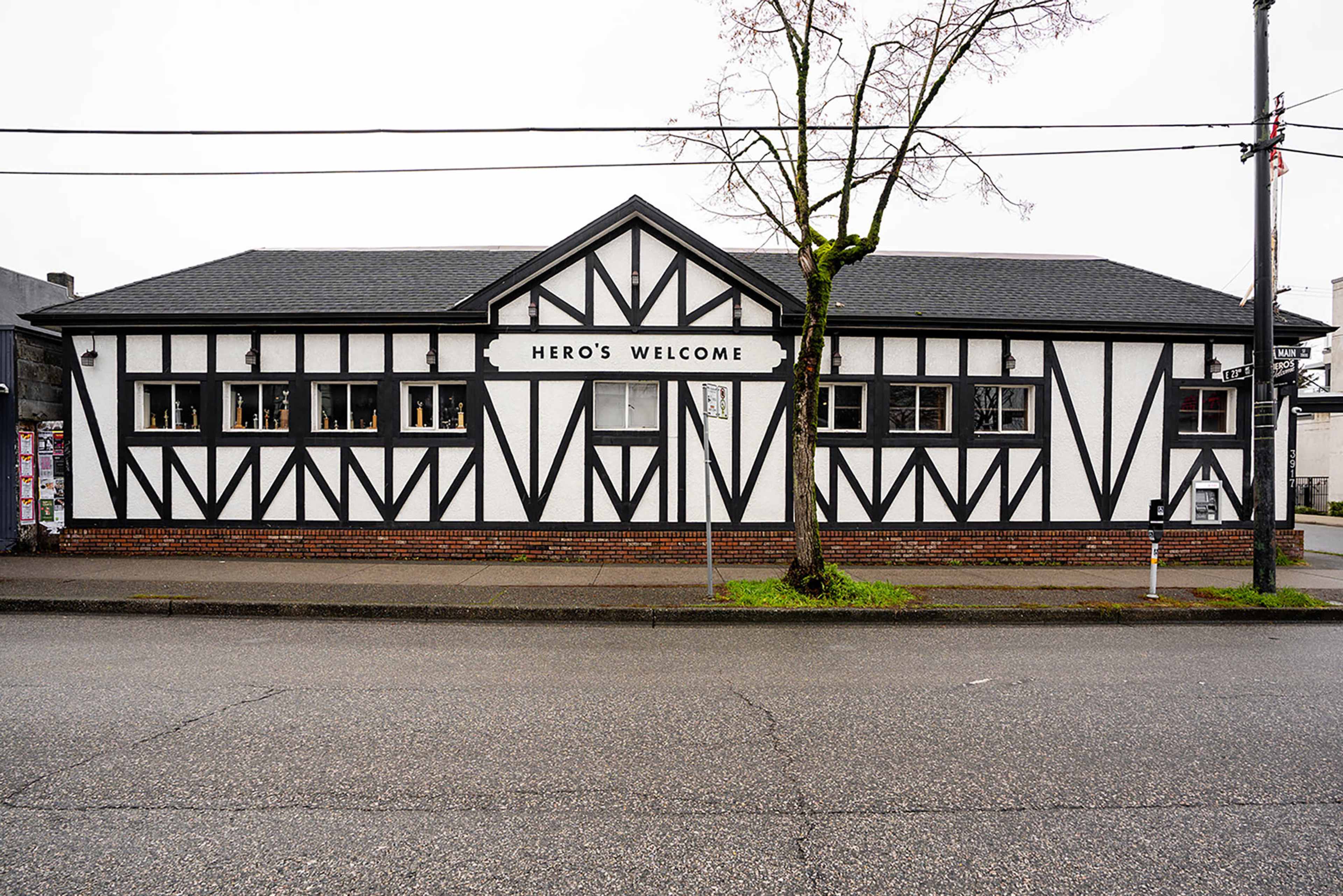 The image shows a distinctively designed building with a black and white half-timbered facade and a sign that reads "Hero's Welcome."