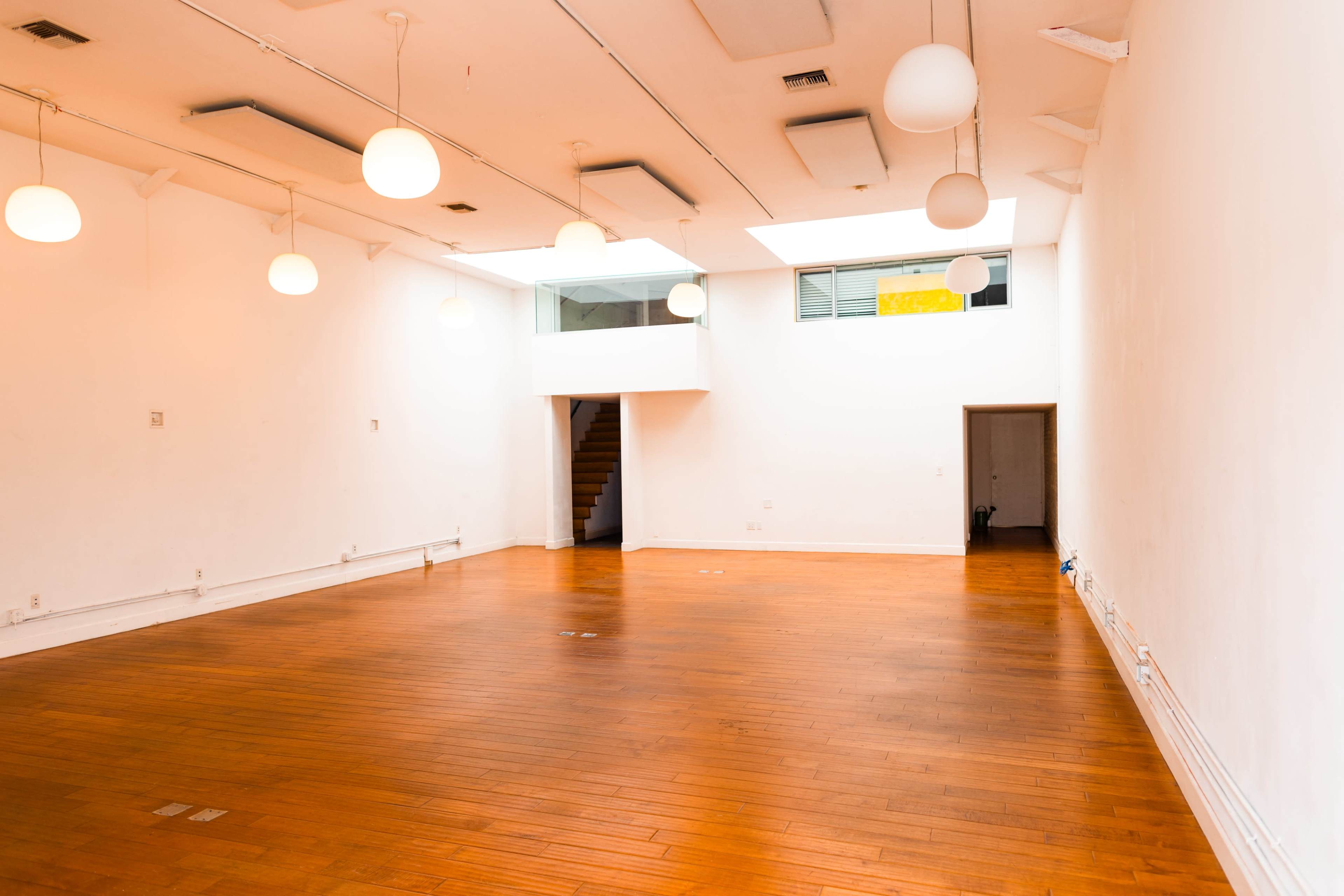 The image shows an empty, well-lit room with wooden flooring and white walls, featuring hanging light fixtures and a staircase in the background.