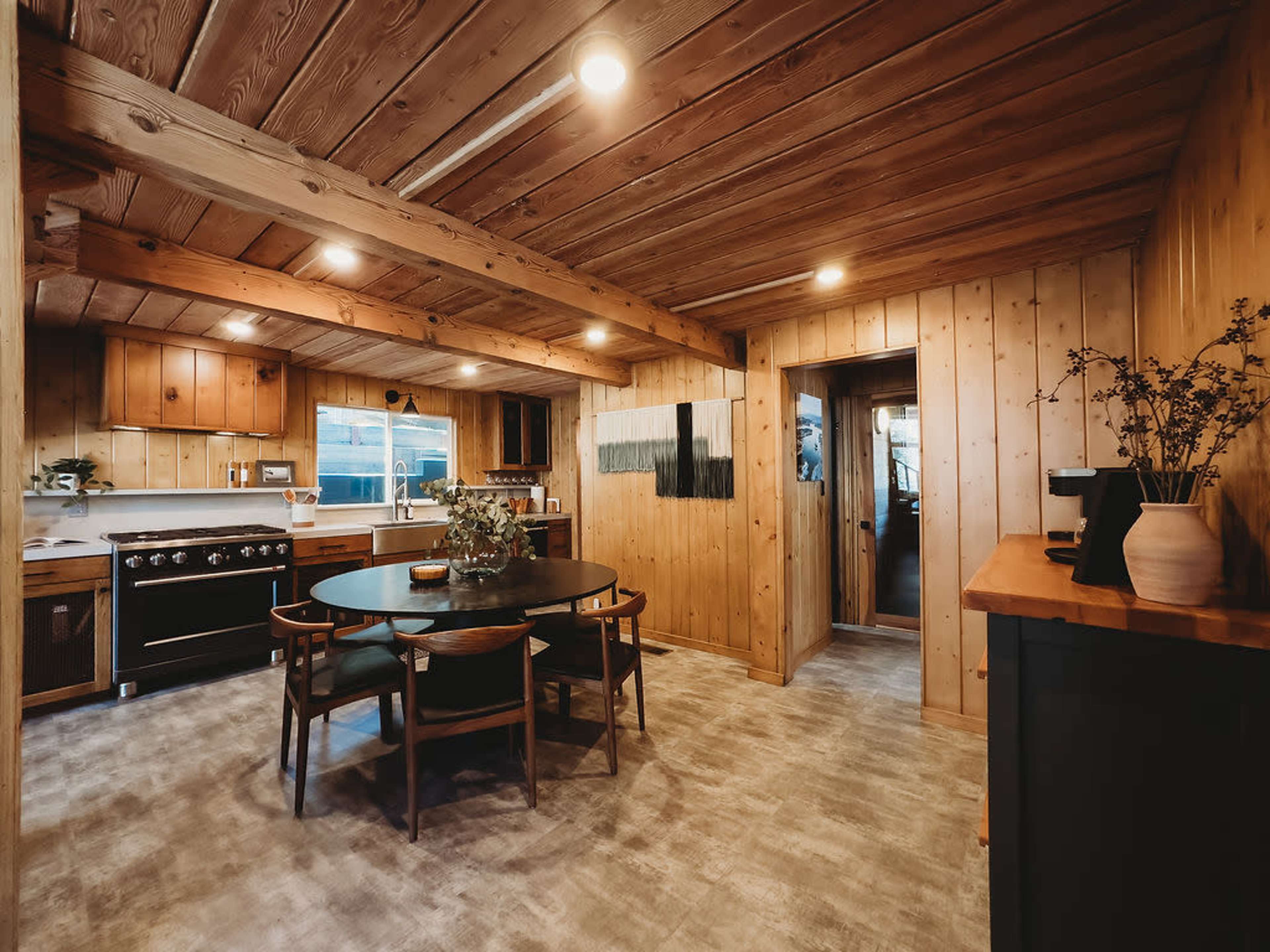 The image shows a wooden-finished kitchen and dining area featuring a round dining table, a large stove, and a doorway leading to another room.