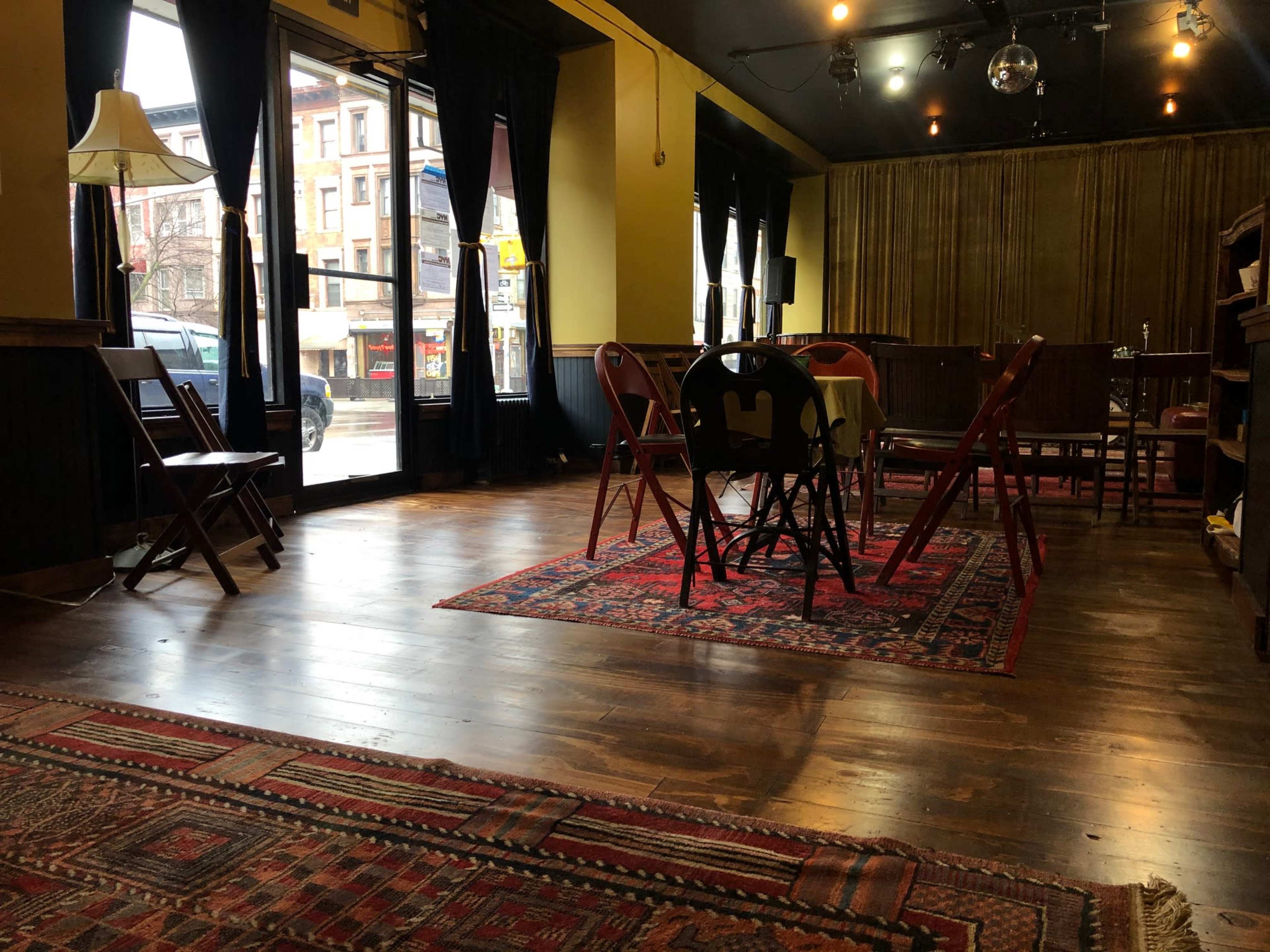 A spacious, empty room with wooden floors, a decorative rug, multiple chairs and tables, and large windows facing a street.